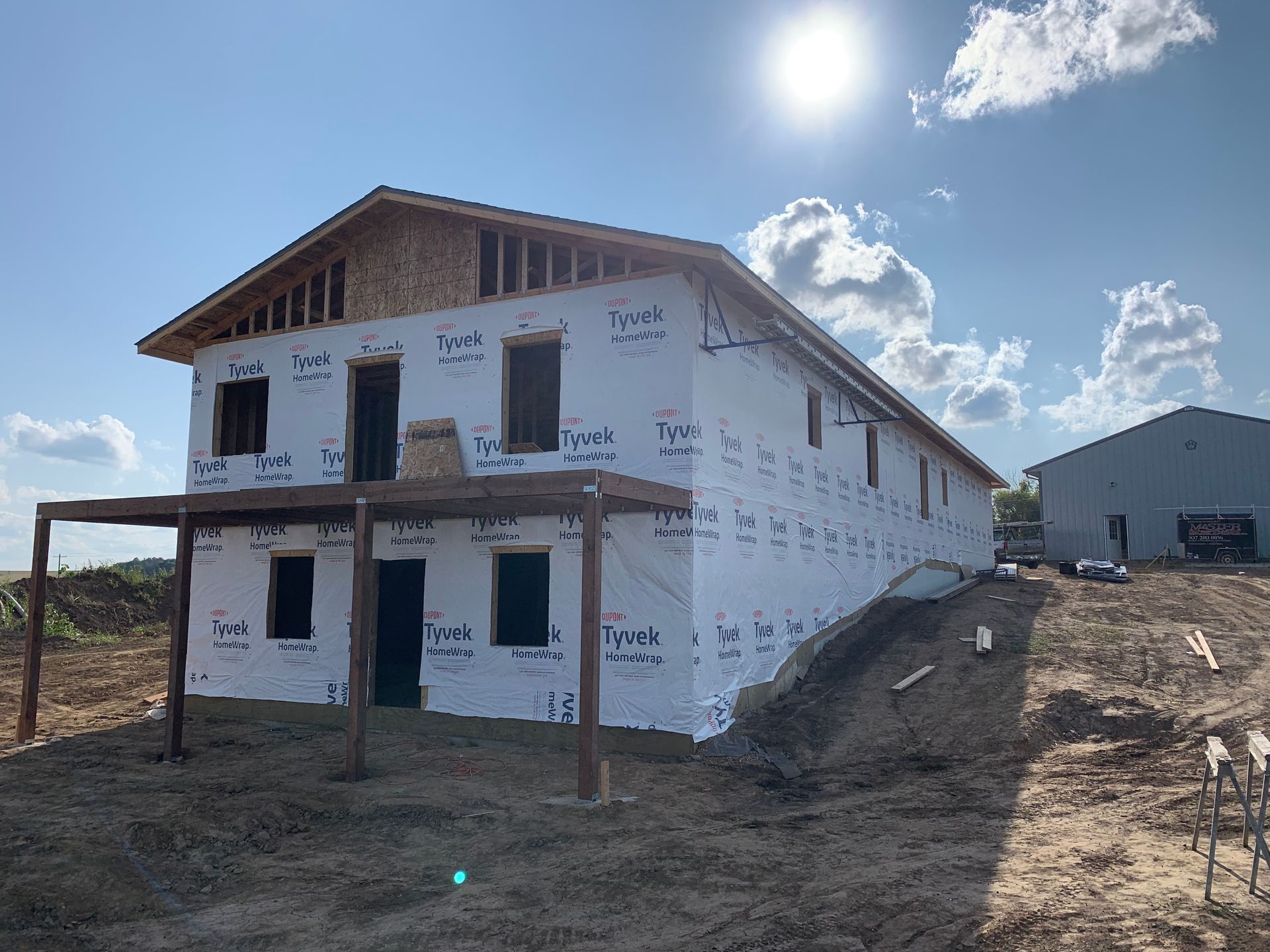 Two-story house under construction, wrapped in white, with a sunlit blue sky background.