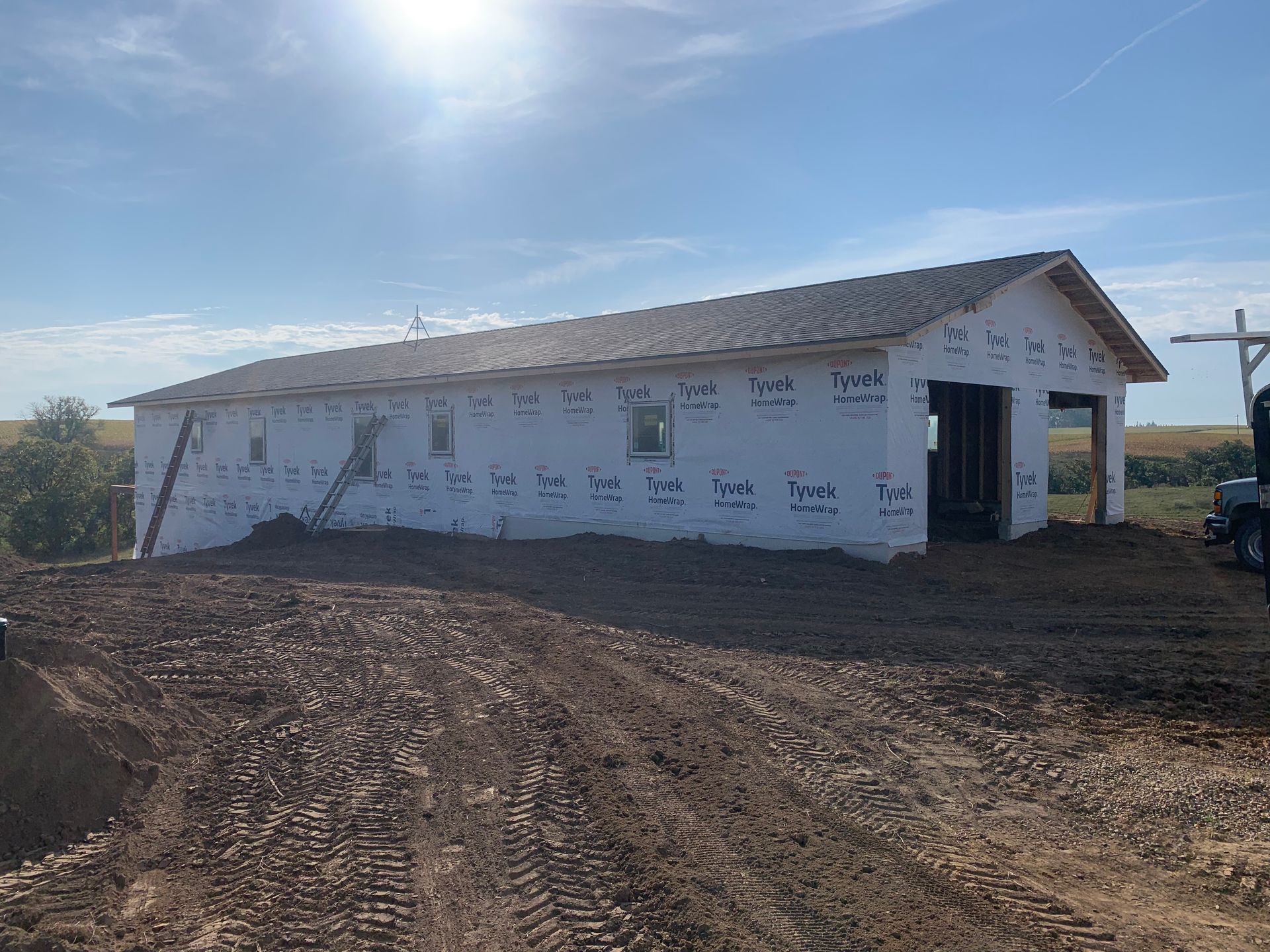 Building under construction, wrapped in white, with roof and two garage door openings; dirt foreground.