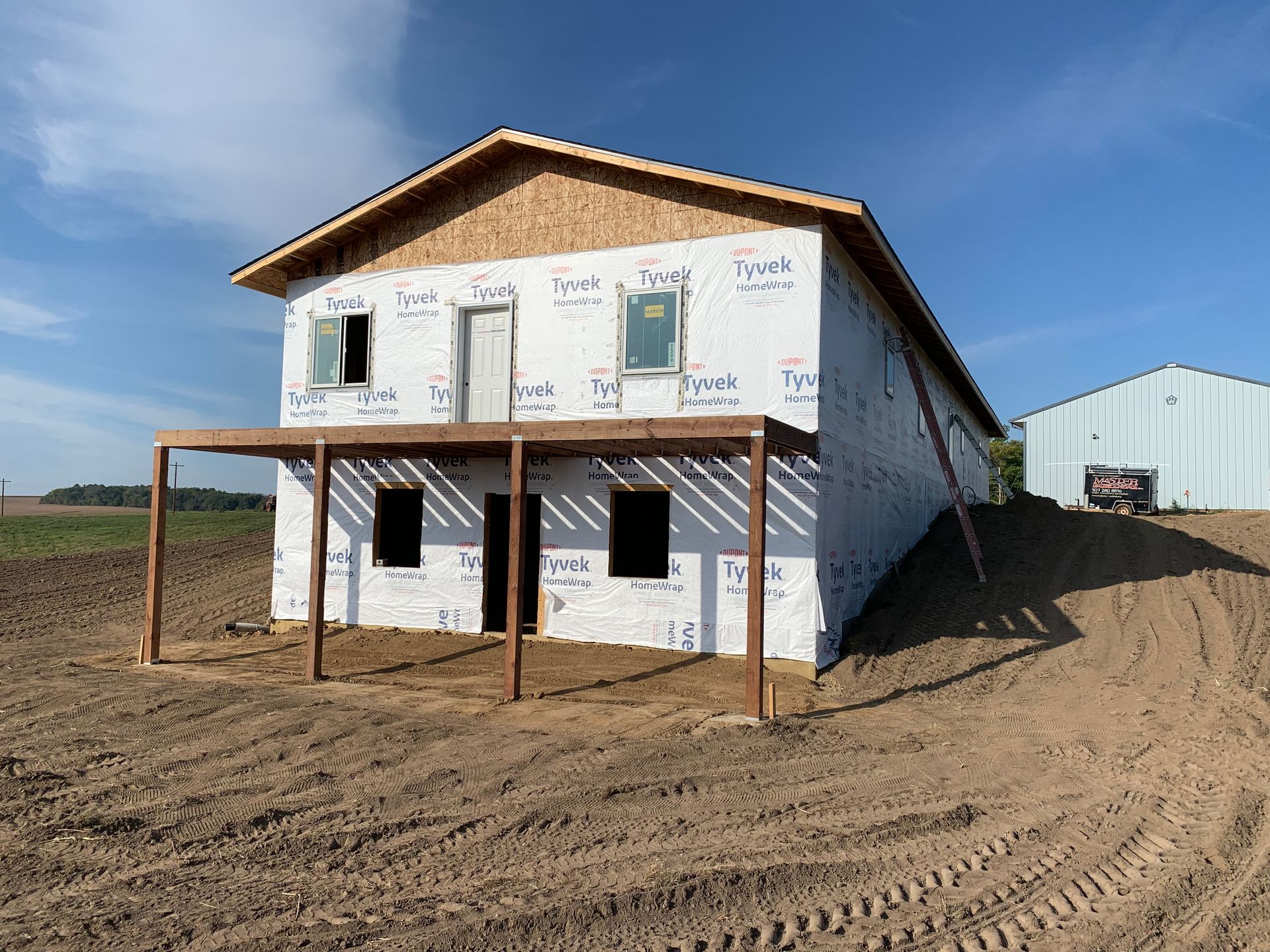 Two-story house under construction, wrapped in white, with unfinished porch. Sunny day, rural setting.