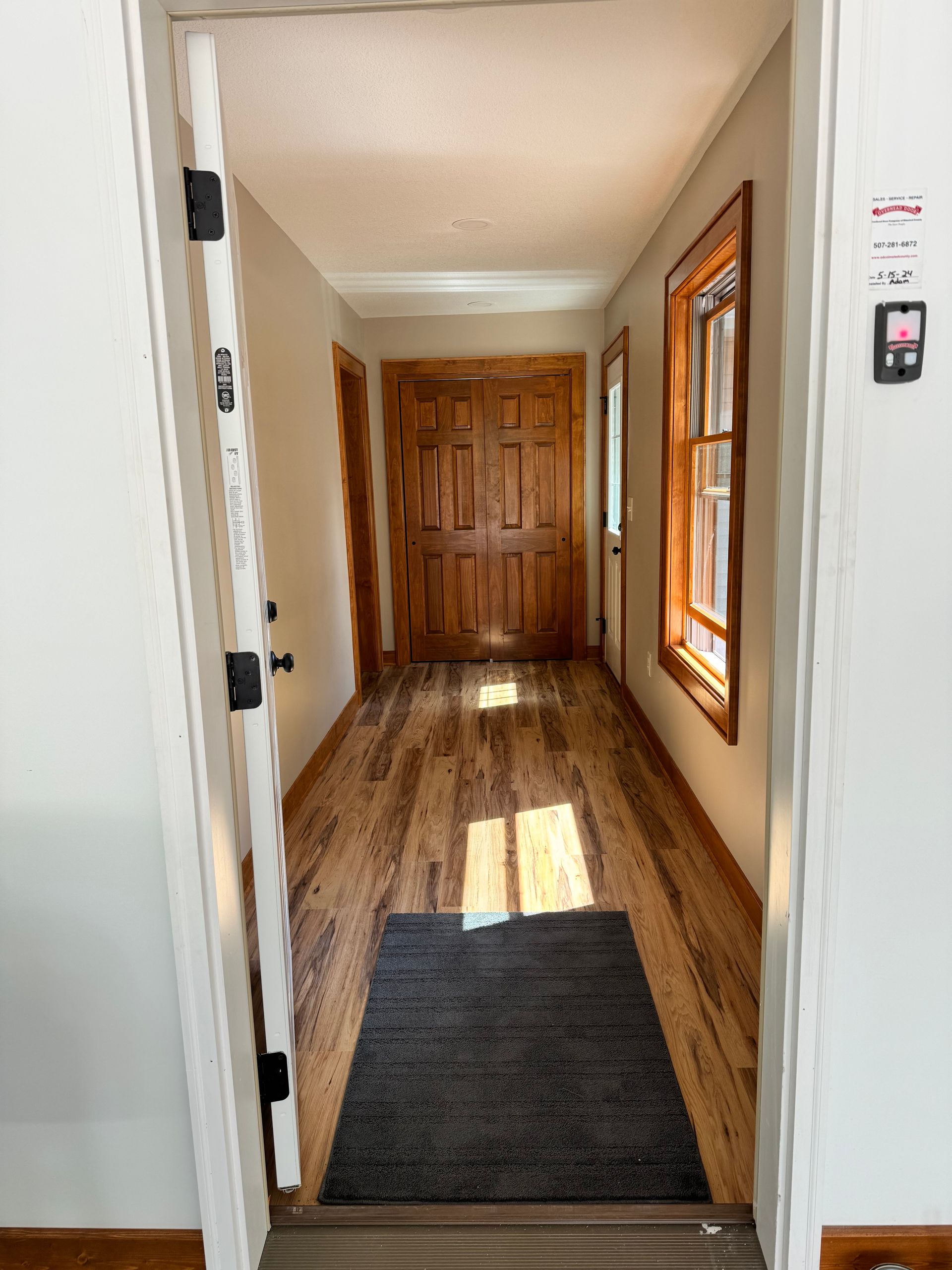 Hallway with hardwood floors, wooden doors, and a dark rug. Sunlight streams through a window.