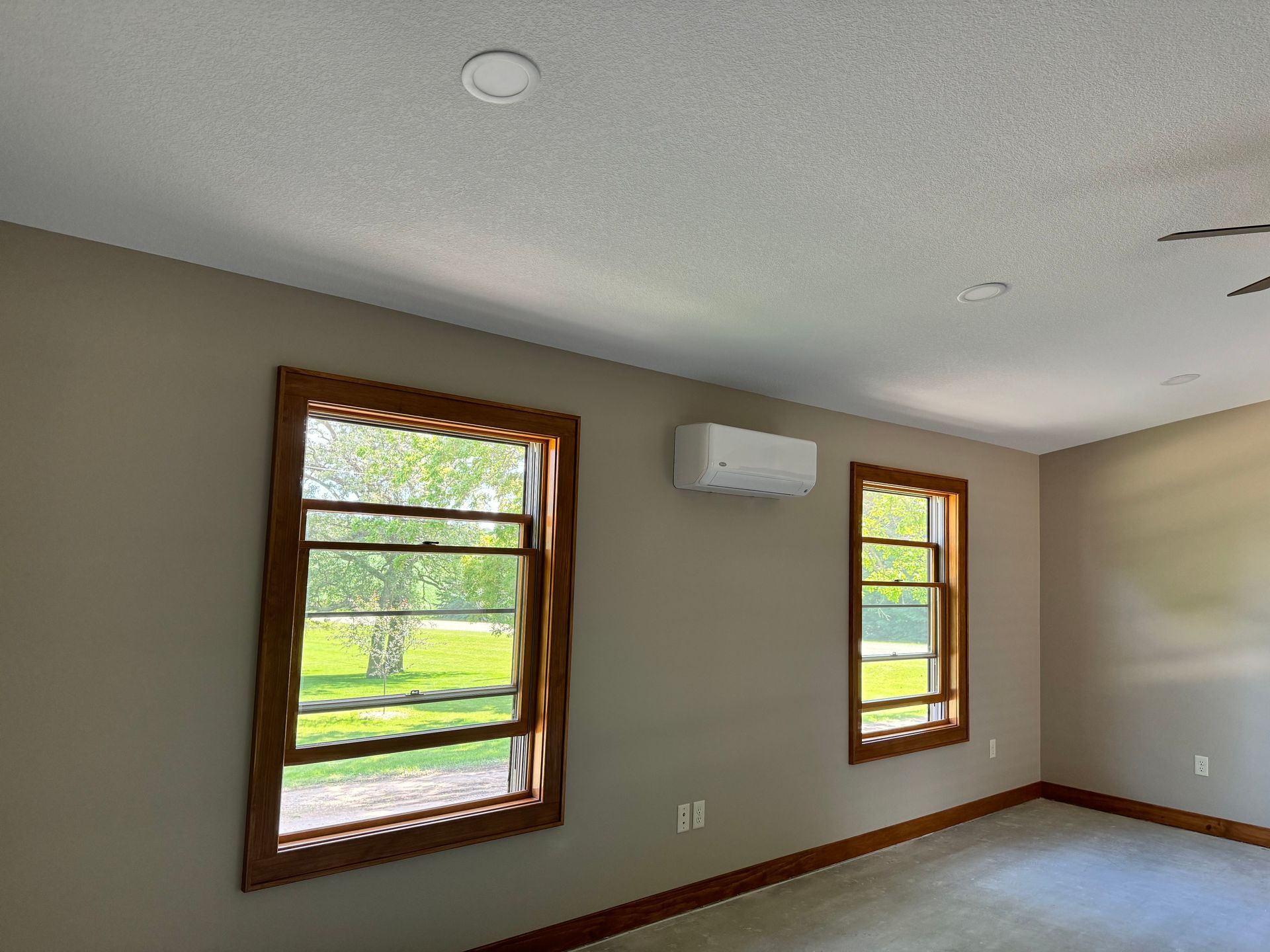 A room with two windows framed in brown wood, a white air conditioner, and beige walls.