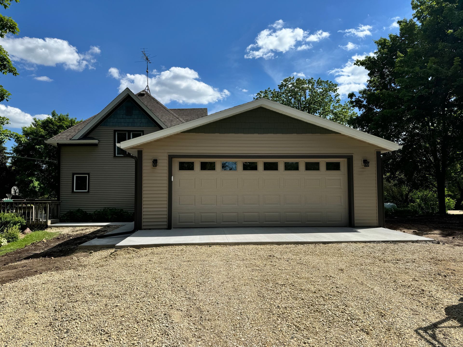 Beige garage with tan door and blue roof, in front of a house with gravel driveway, under a blue sky.