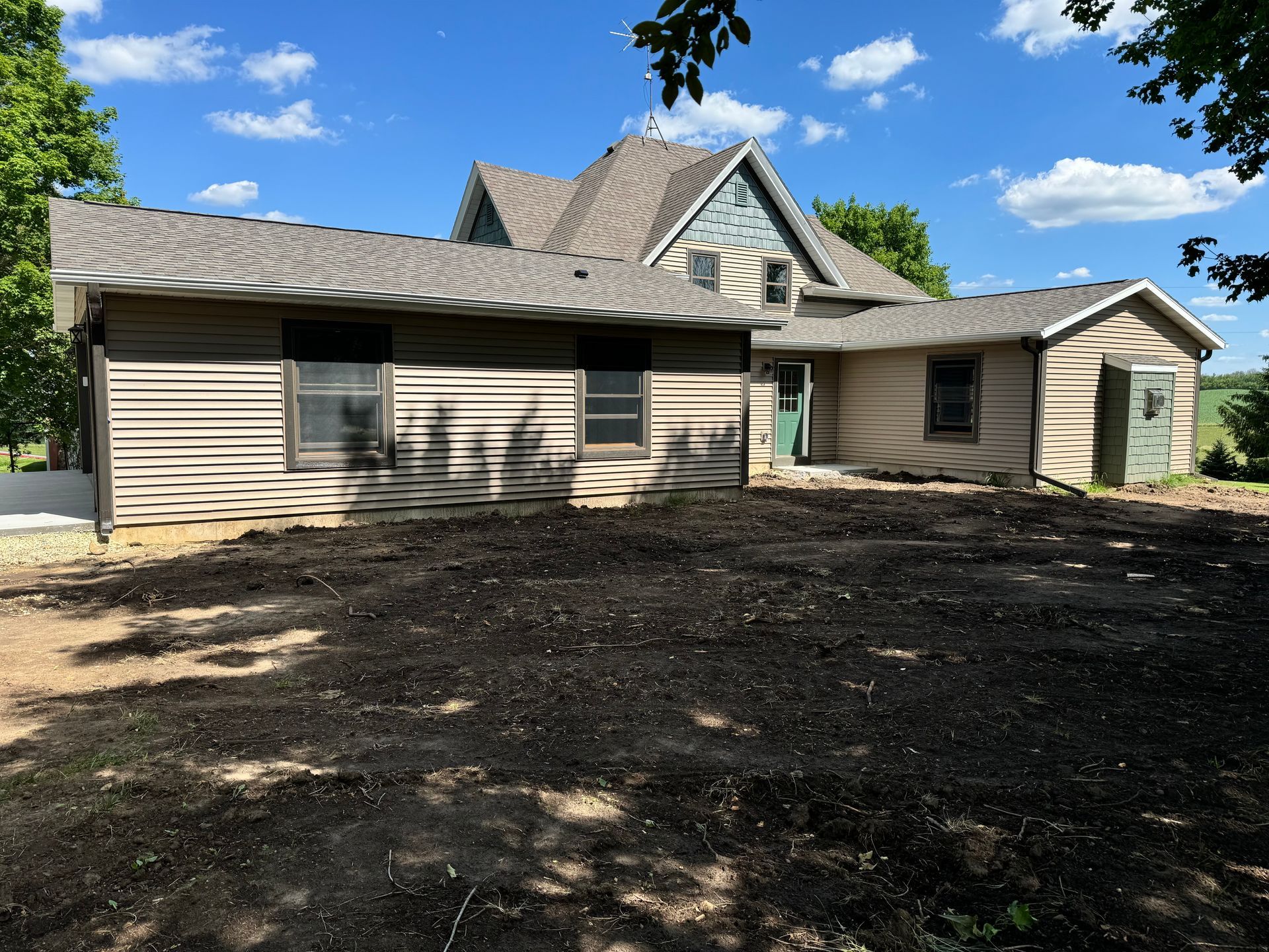 Backyard view of a house with a brown roof and siding, dark soil, and blue sky.