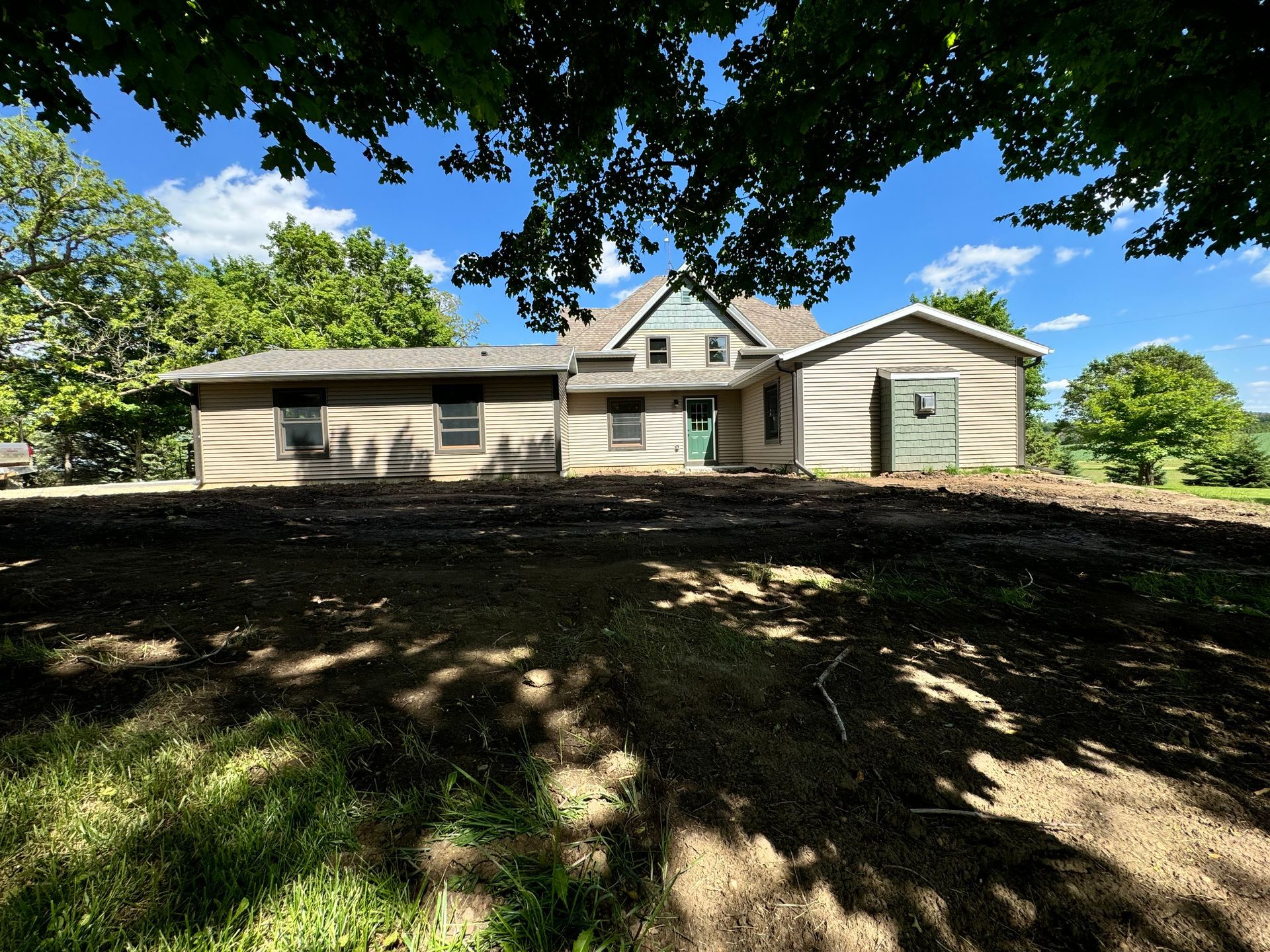 Old farmhouse under a blue sky, viewed from a grassy foreground shaded by trees.