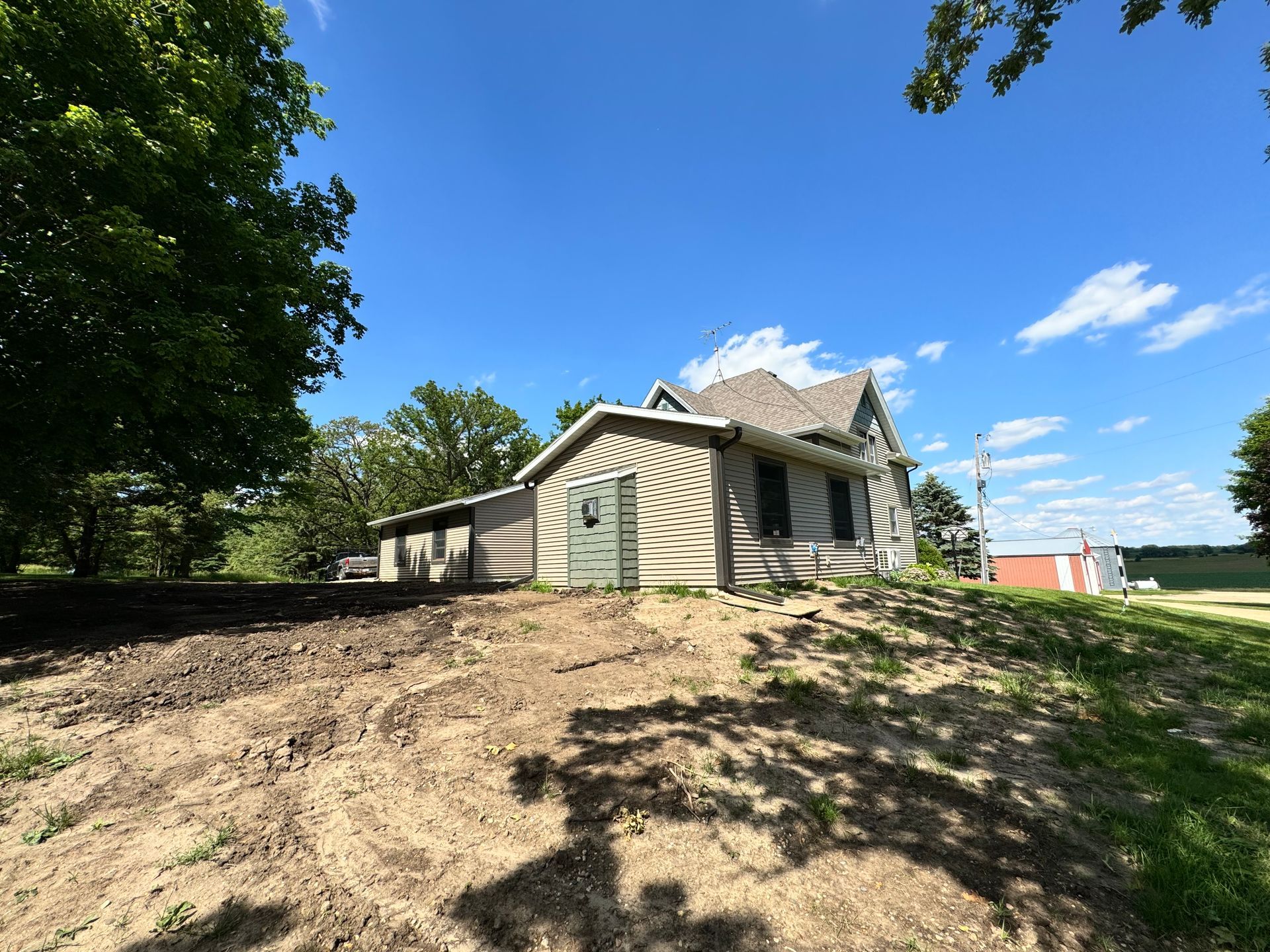 Farmhouse on a slight hill with a shed, trees, and blue sky.