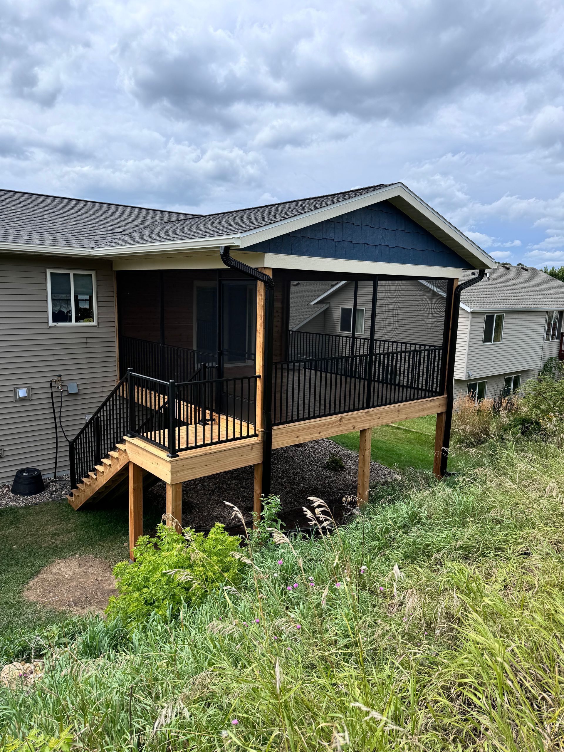 Screened-in porch attached to a house. Black railing, wooden deck, and steps. Green vegetation in the foreground.