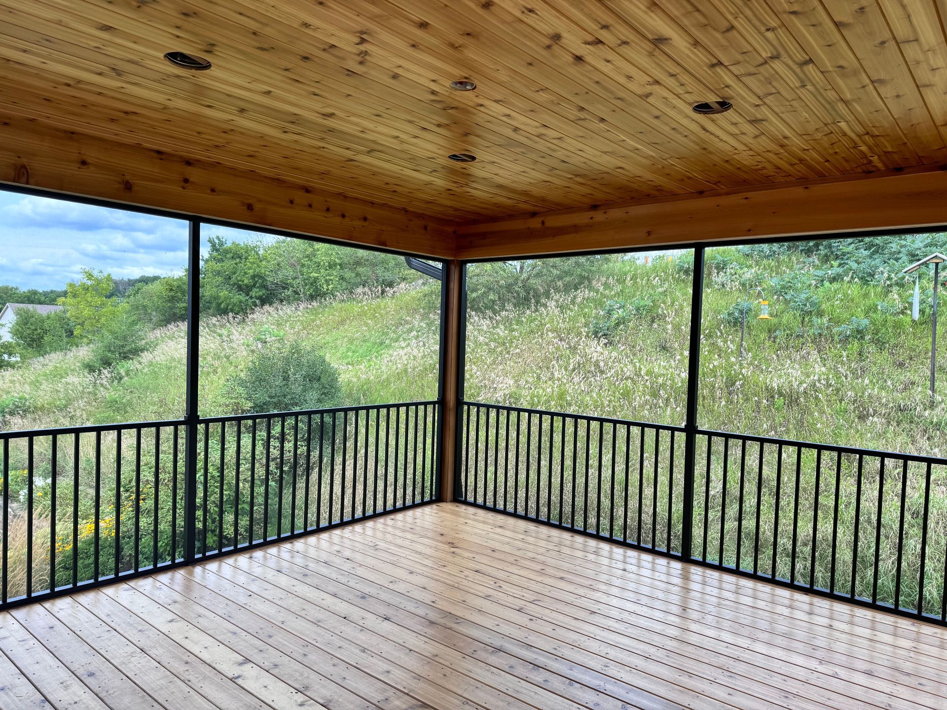 Screened porch with wood ceiling and floor, black railings, overlooking a grassy field.