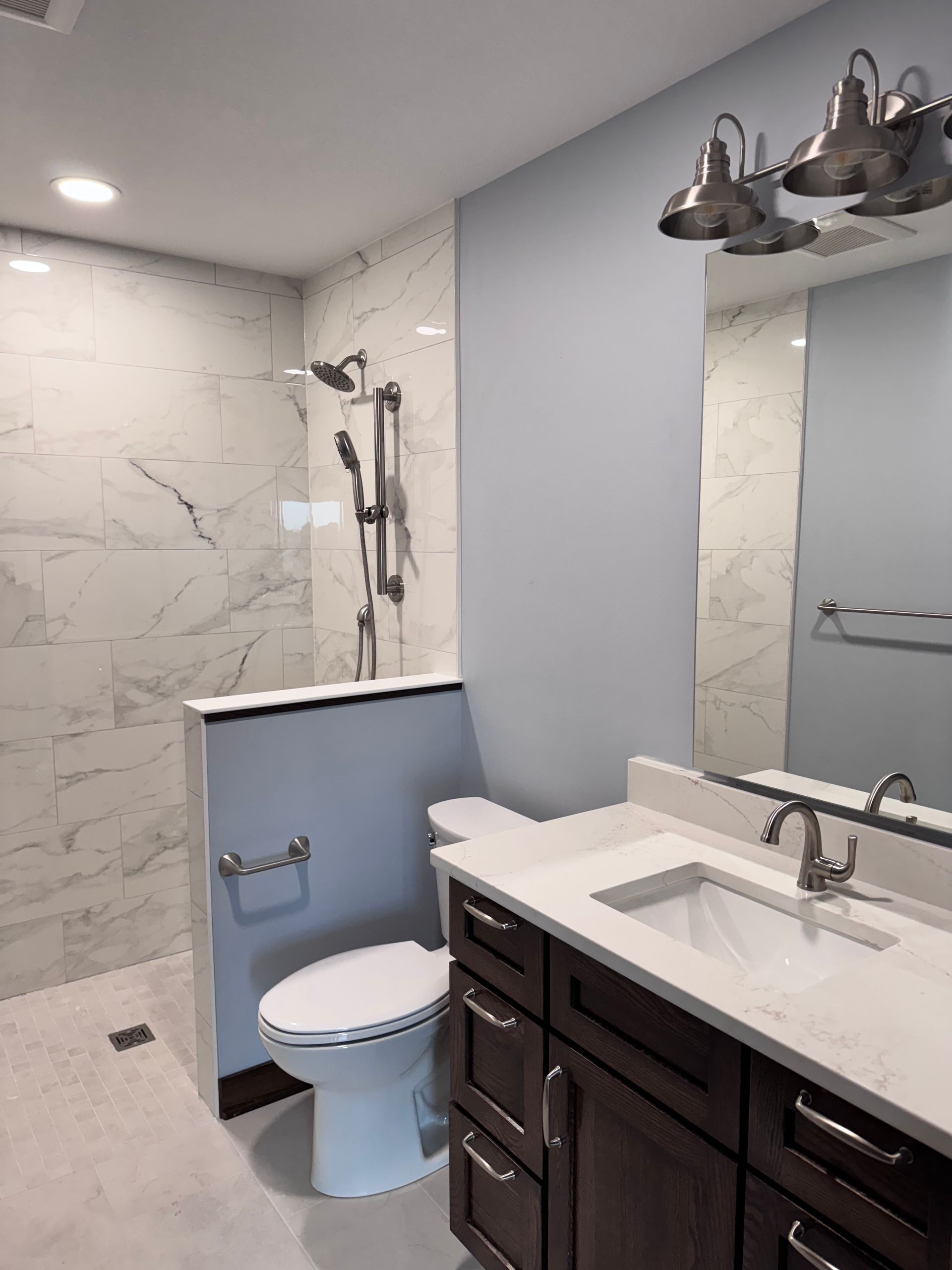 Modern bathroom with marble-tiled shower, toilet, vanity, and overhead lights. Light blue walls contrast dark cabinets.