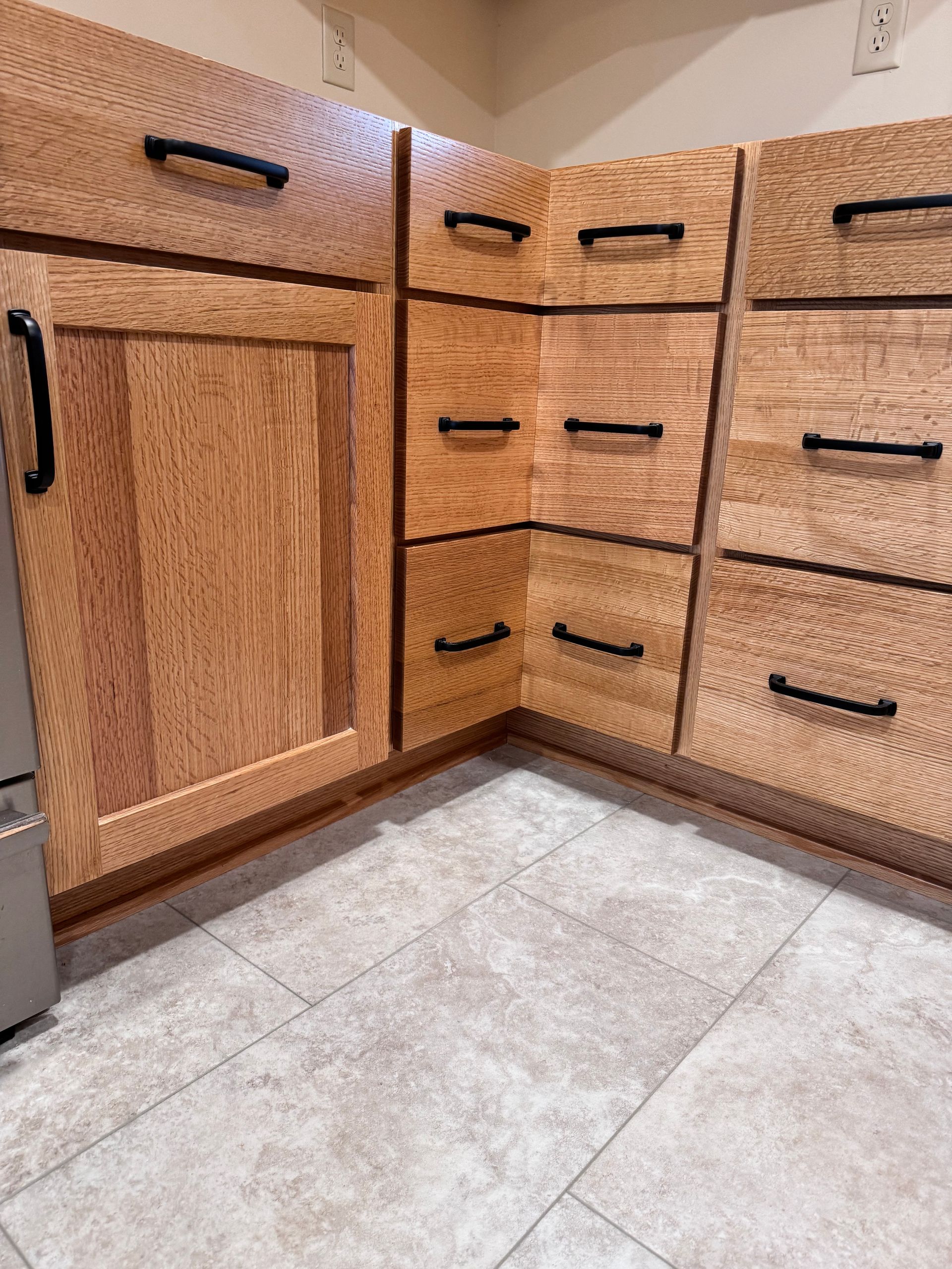 Corner kitchen cabinets in light wood with black handles, set on tan tile flooring.