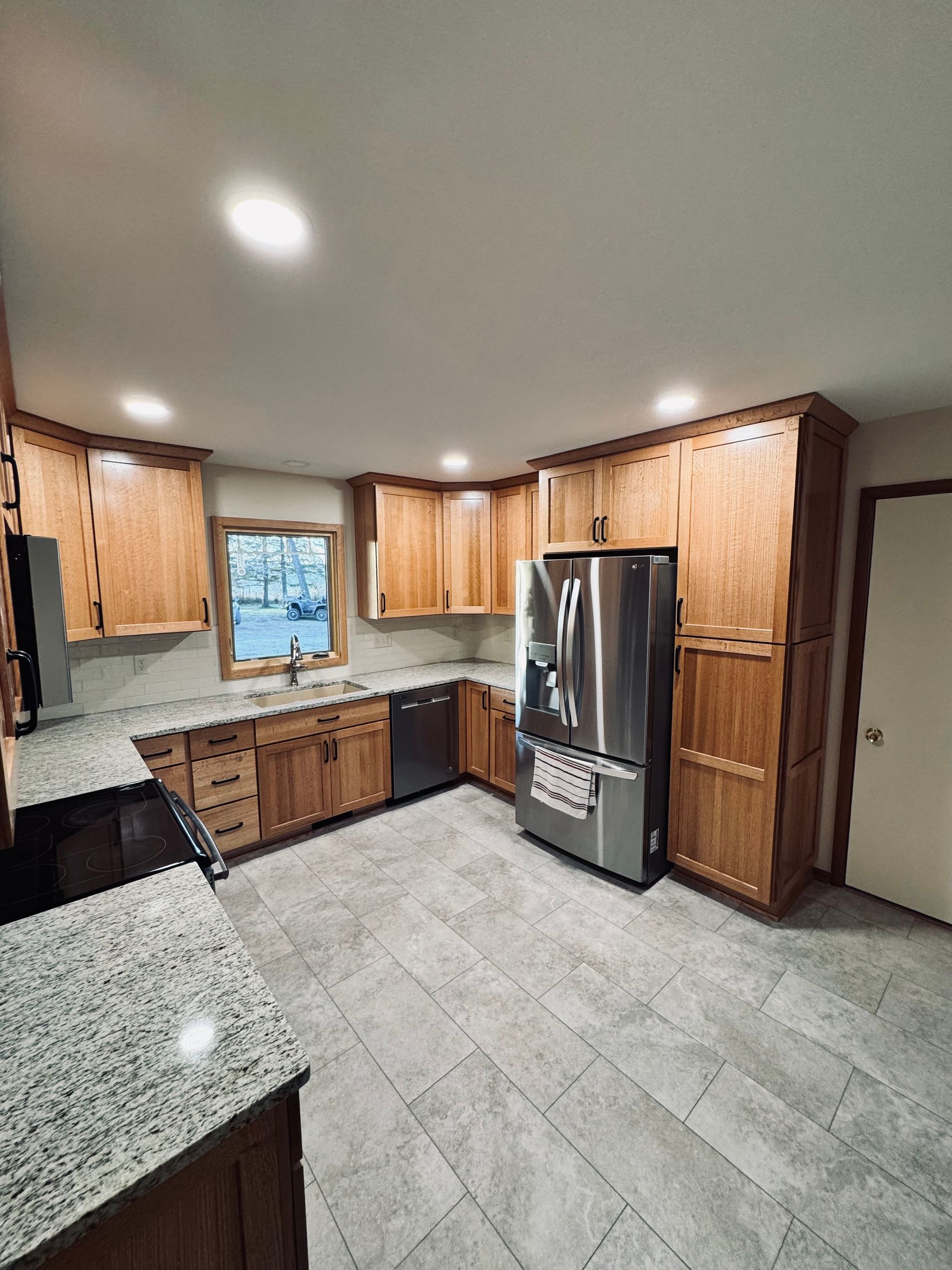 A modern kitchen with light wood cabinets, stainless steel appliances, gray countertops, and tile floors.