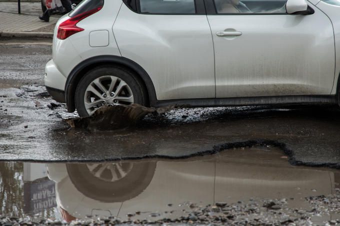 Back-end of a small white vehicle hitting a deep pothole while driving on a street