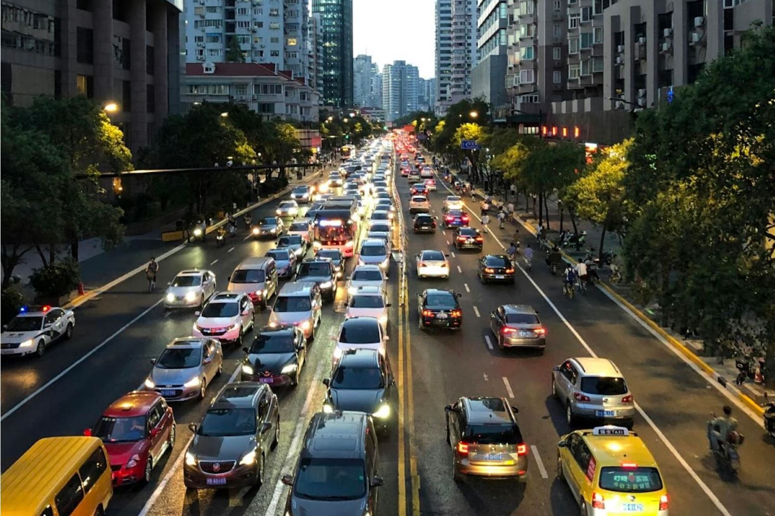A congested city street at dusk filled with cars in multiple lanes, surrounded by tall buildings and trees.