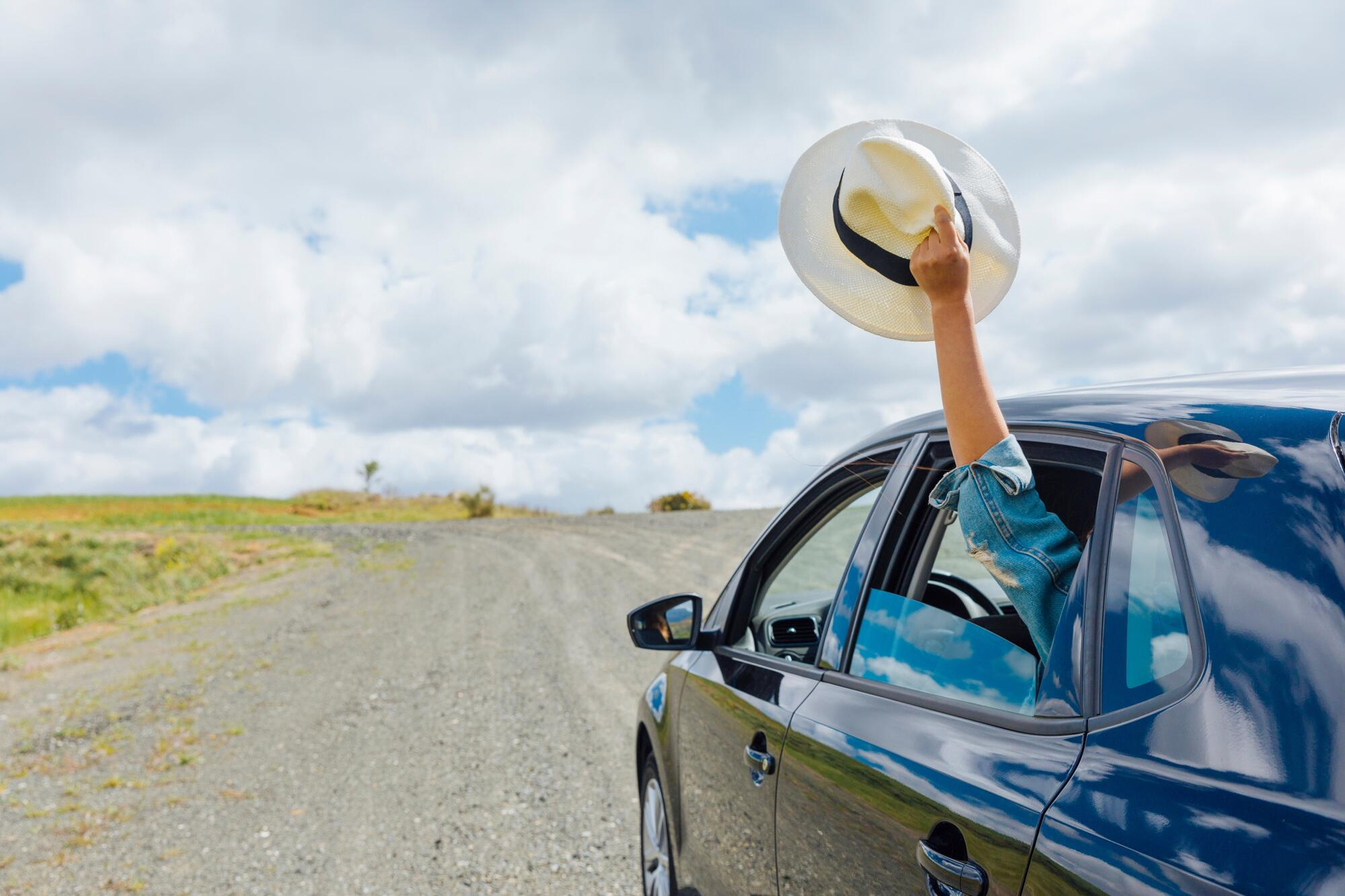 Person in a black car raises a hat out the window on a gravel road