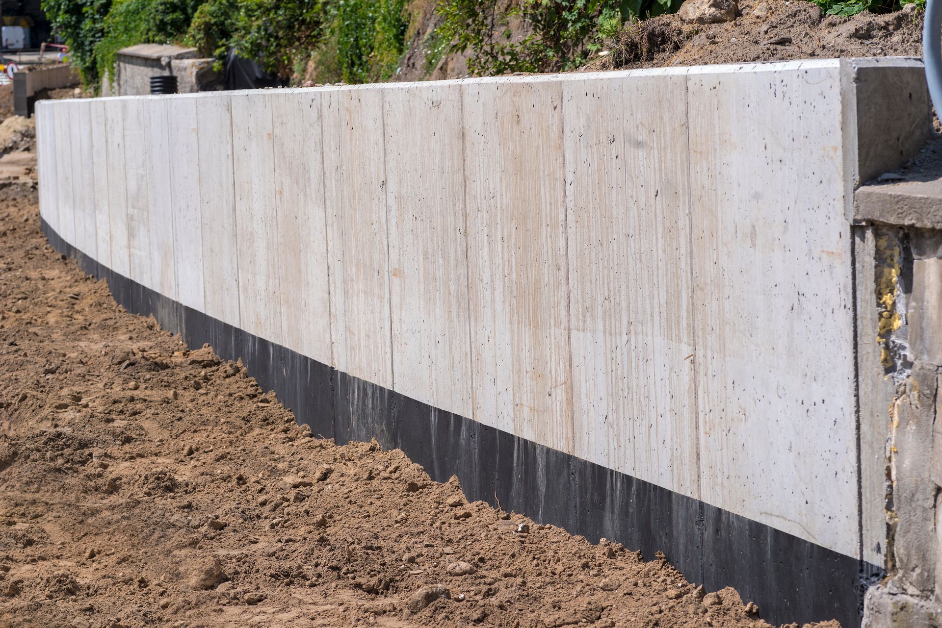 A newly constructed concrete retaining wall with a black waterproofing membrane along the base, set in a dirt landscape.