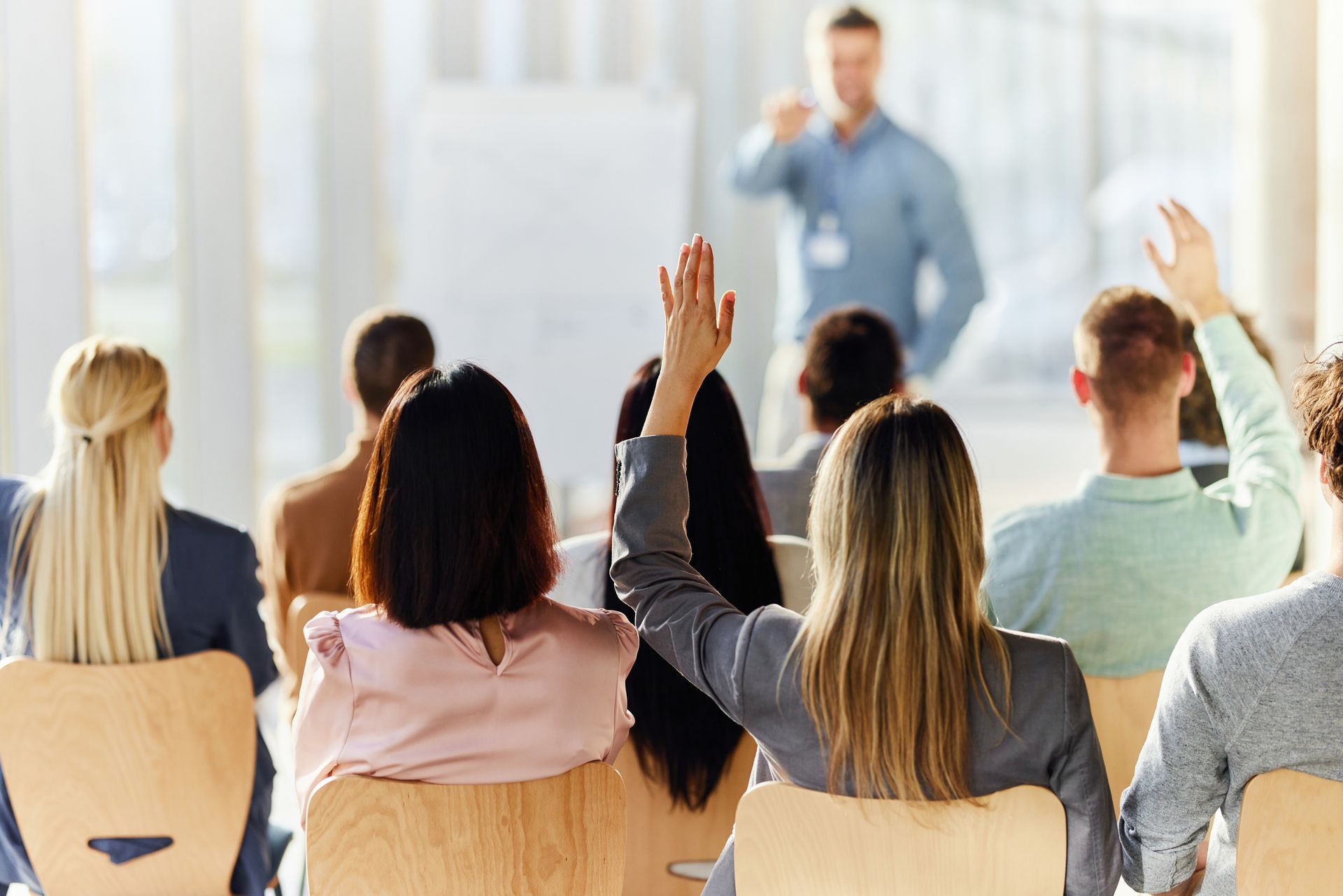 People raising hands in a conference room as a speaker addresses them.