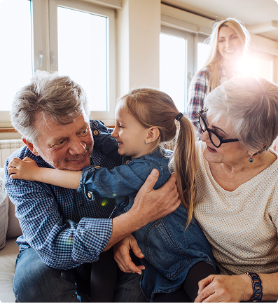 Grandfather hugging a girl, grandmother watches. Another woman stands in the background near a sunny window.