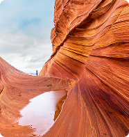 Orange sandstone canyon with a small pool of water reflecting the sky; a person stands in the distance.