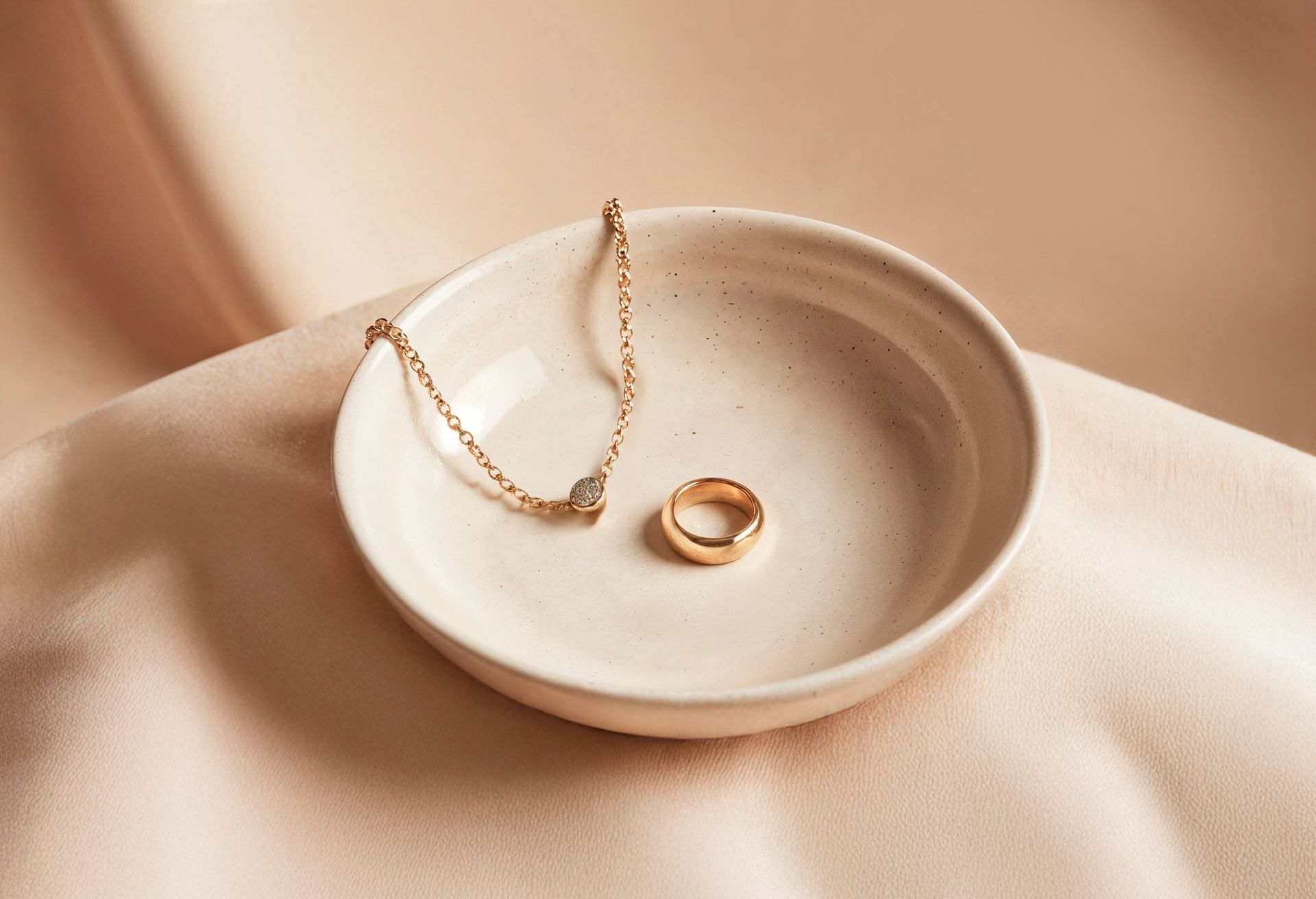 Pequeño cuenco blanco que contiene un anillo de oro y un delicado collar sobre un paño beige drapeado.