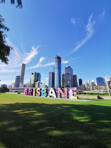 A palavra Brisbane está pintada na grama em frente ao horizonte de uma cidade.