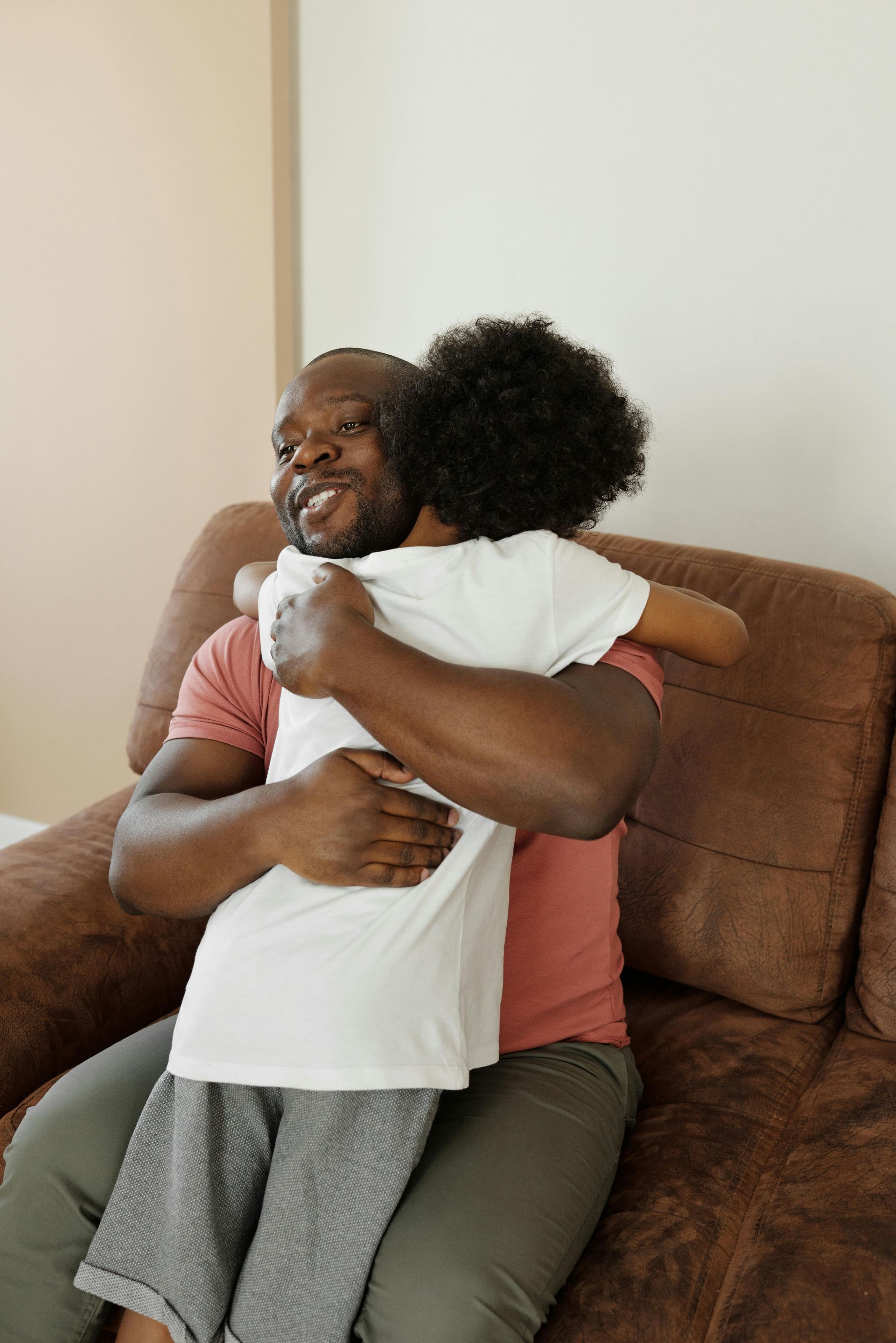 Father and child embrace on a brown couch. The father smiles, wearing a red shirt; the child has curly hair and a white shirt.