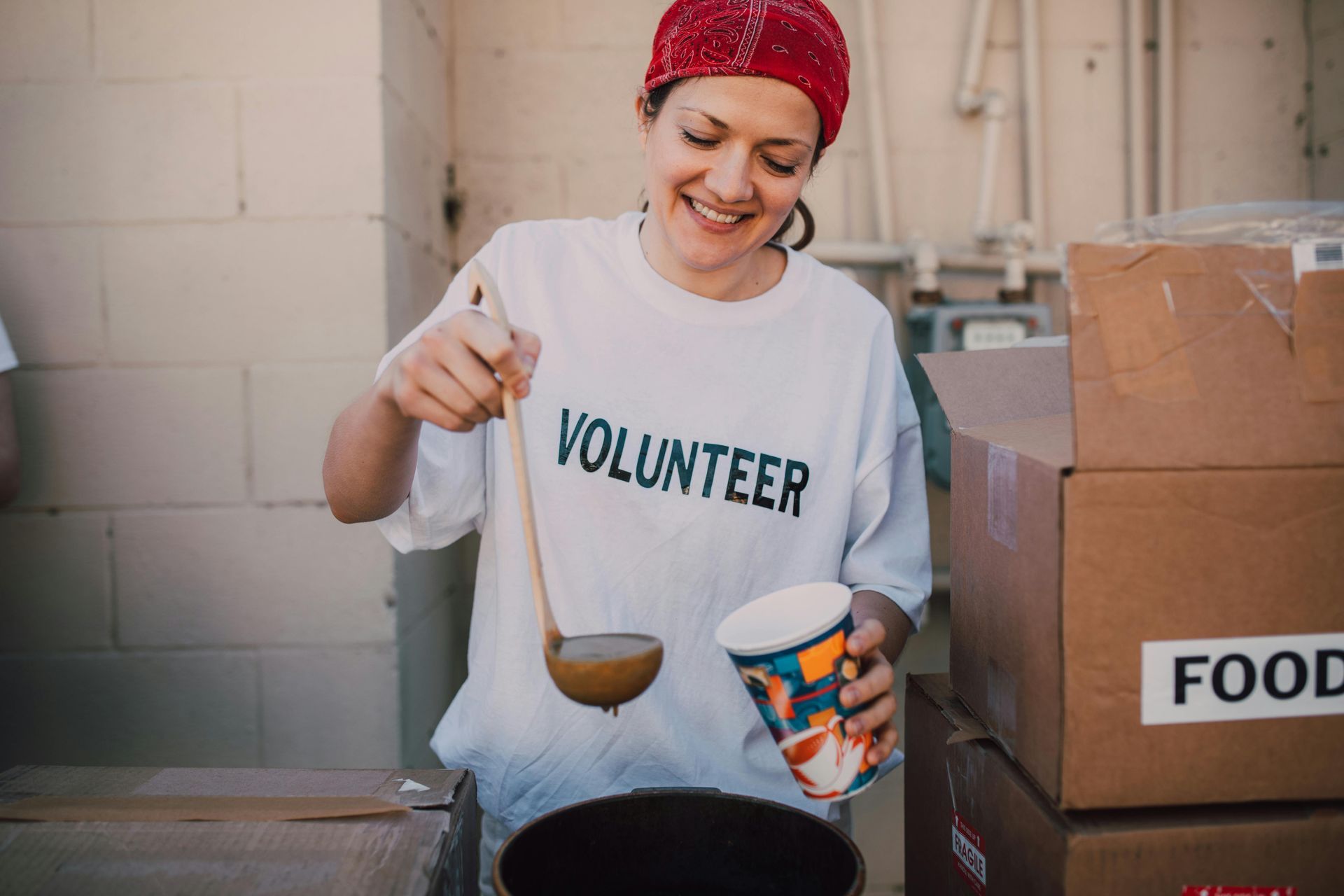 Volunteer ladling soup into a cup. She's smiling, wearing a red bandana and a shirt that says 