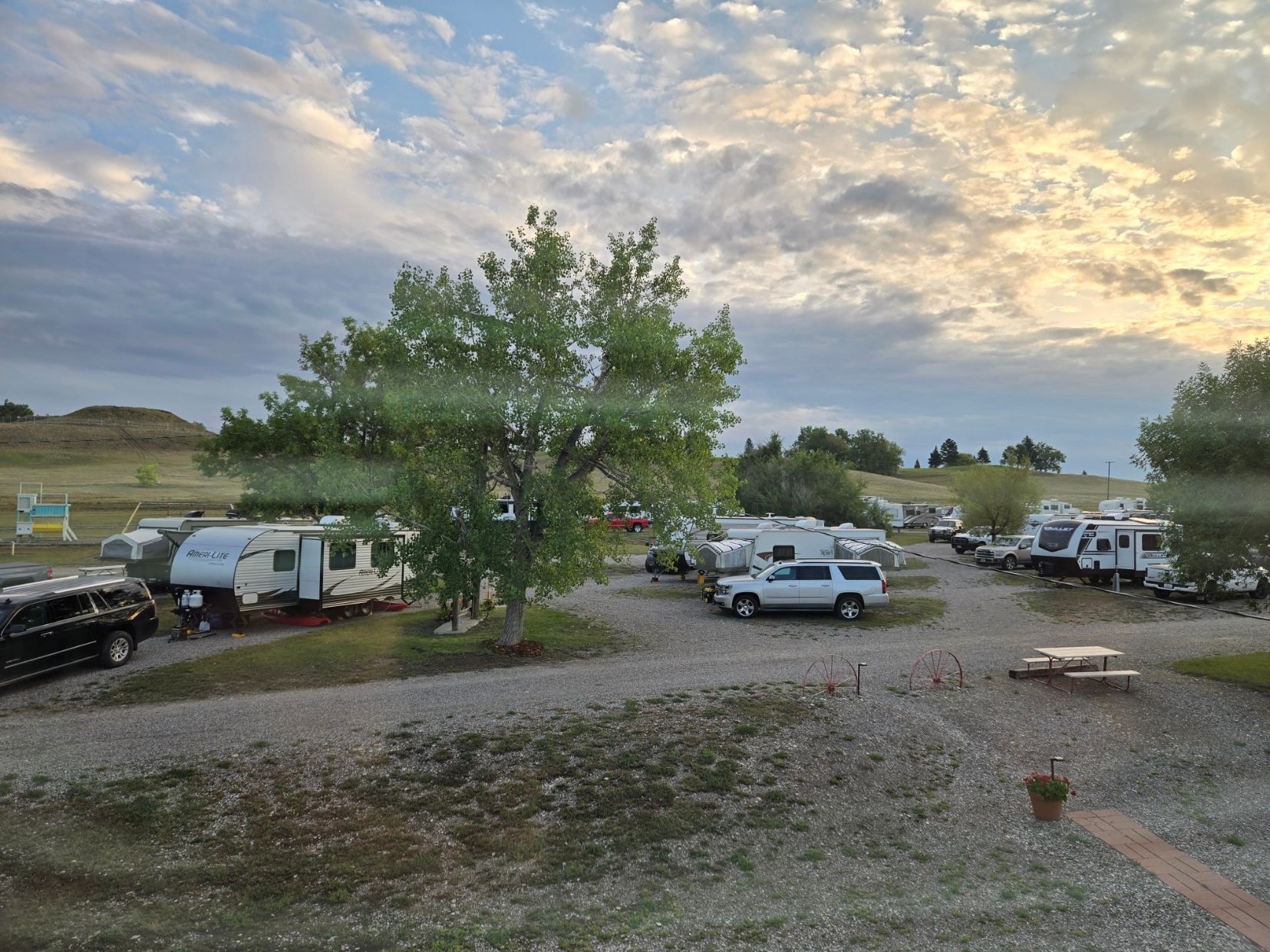 RV park with trees, gravel, and RVs under a cloudy sky.