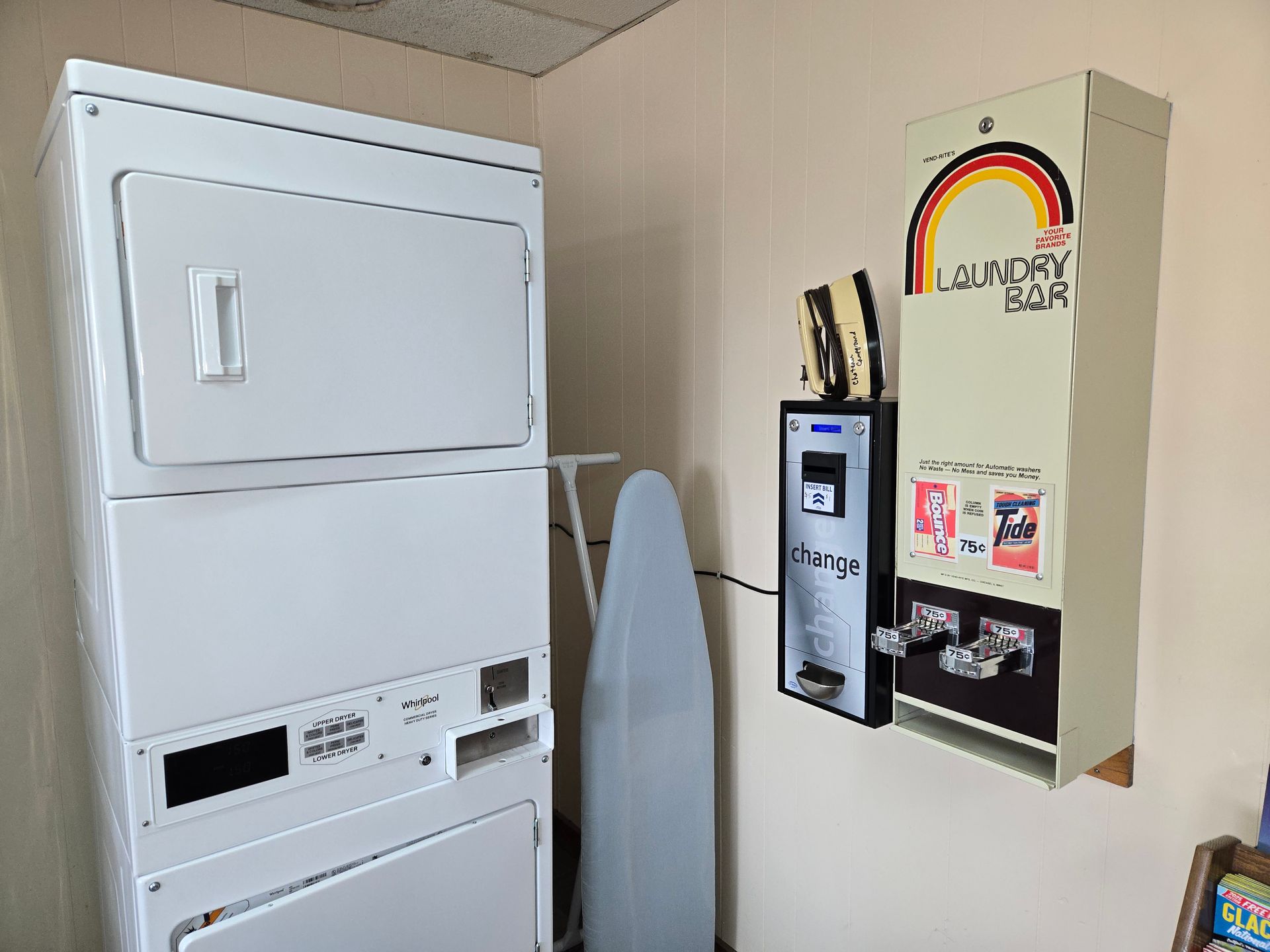 Laundry machines in a room: stacked dryer and washing machine, ironing board, change machine, and detergent vending.