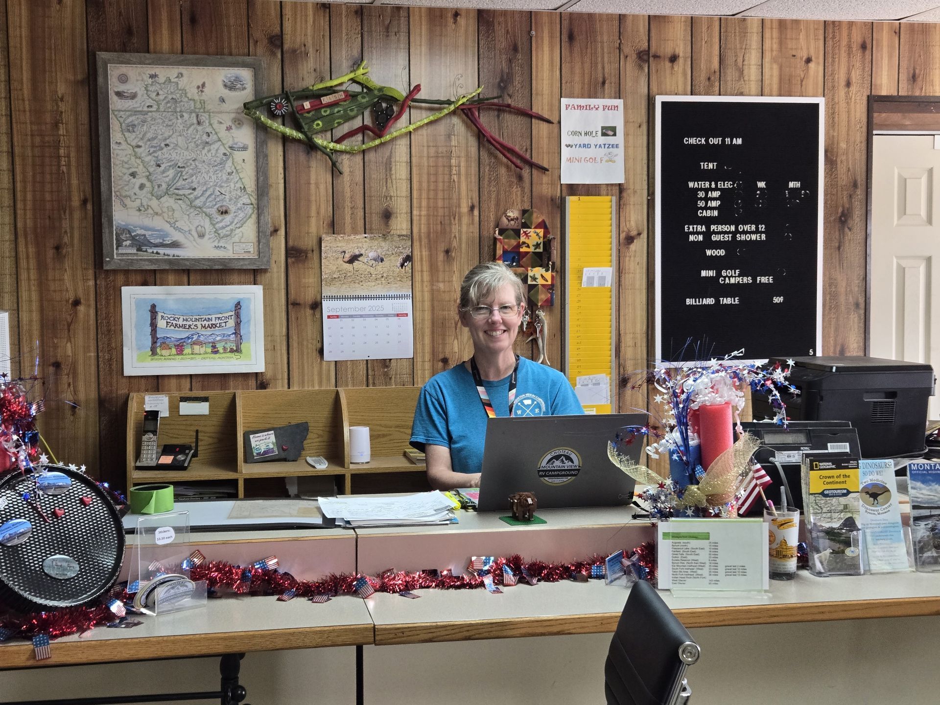 Woman at a desk with a laptop smiles, wooden wall background, decorations, map, and bulletin board.