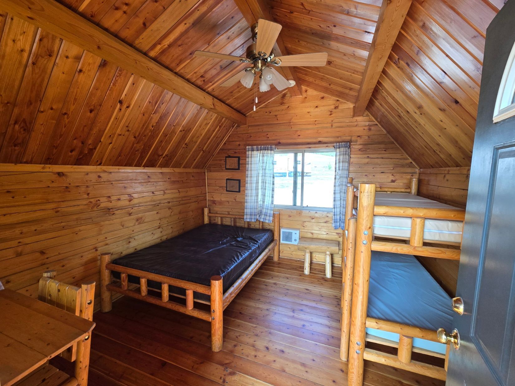 Wooden cabin interior with bed, bunk beds, window, and ceiling fan.