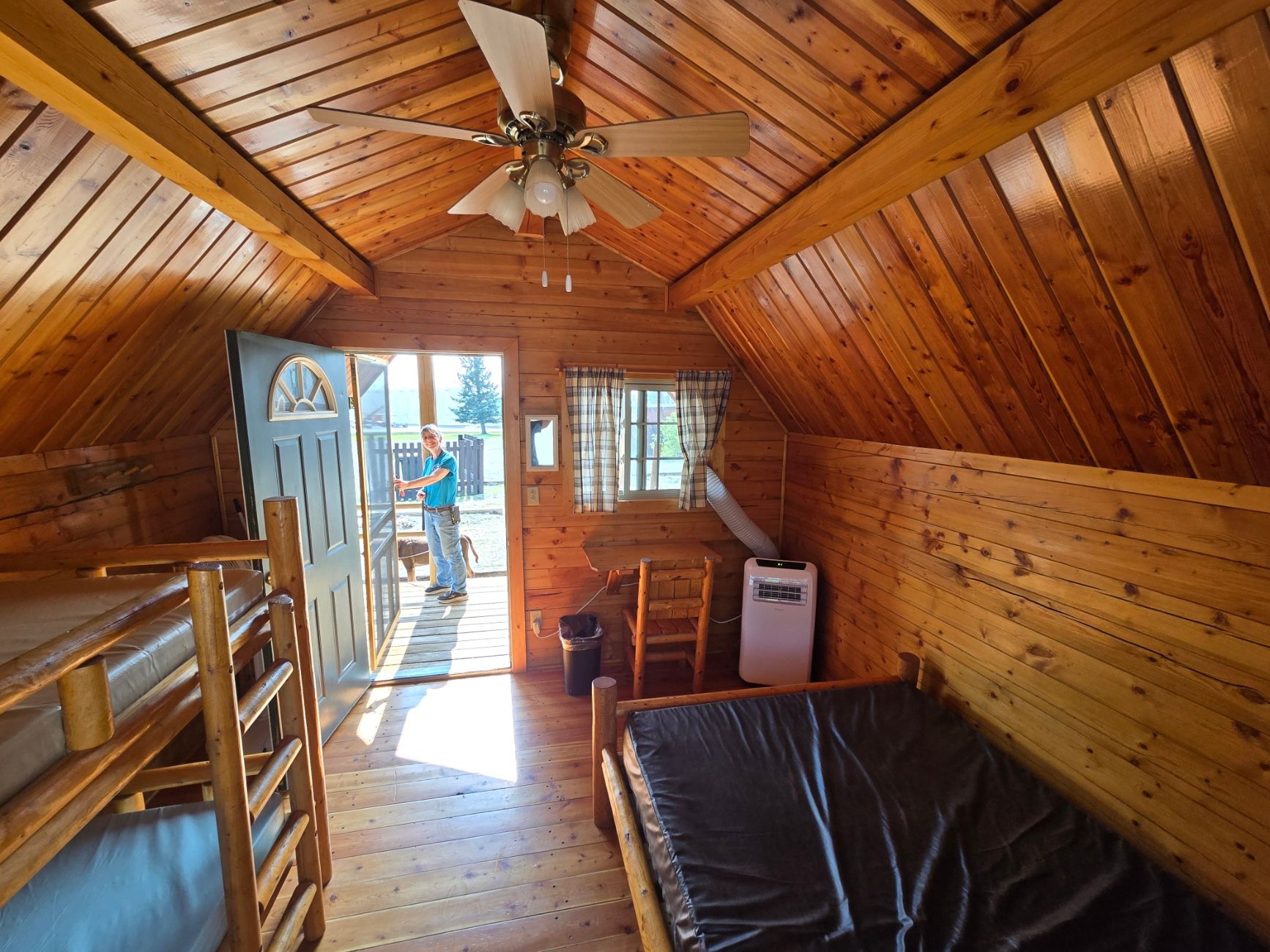 Interior of a cabin with wooden walls, bunk beds, and a single bed. A person stands in the doorway.