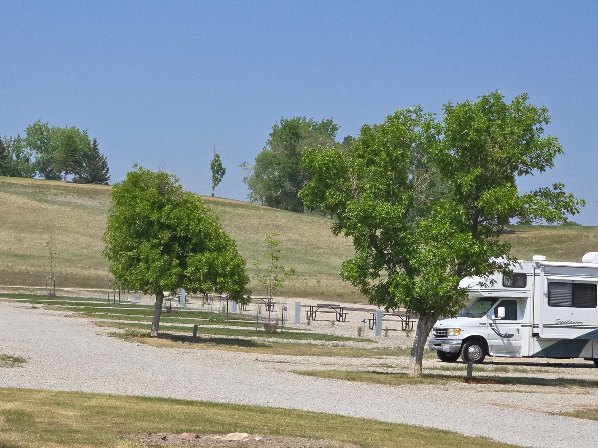RV campsite with gravel roads, trees, and a grassy hill under a blue sky.