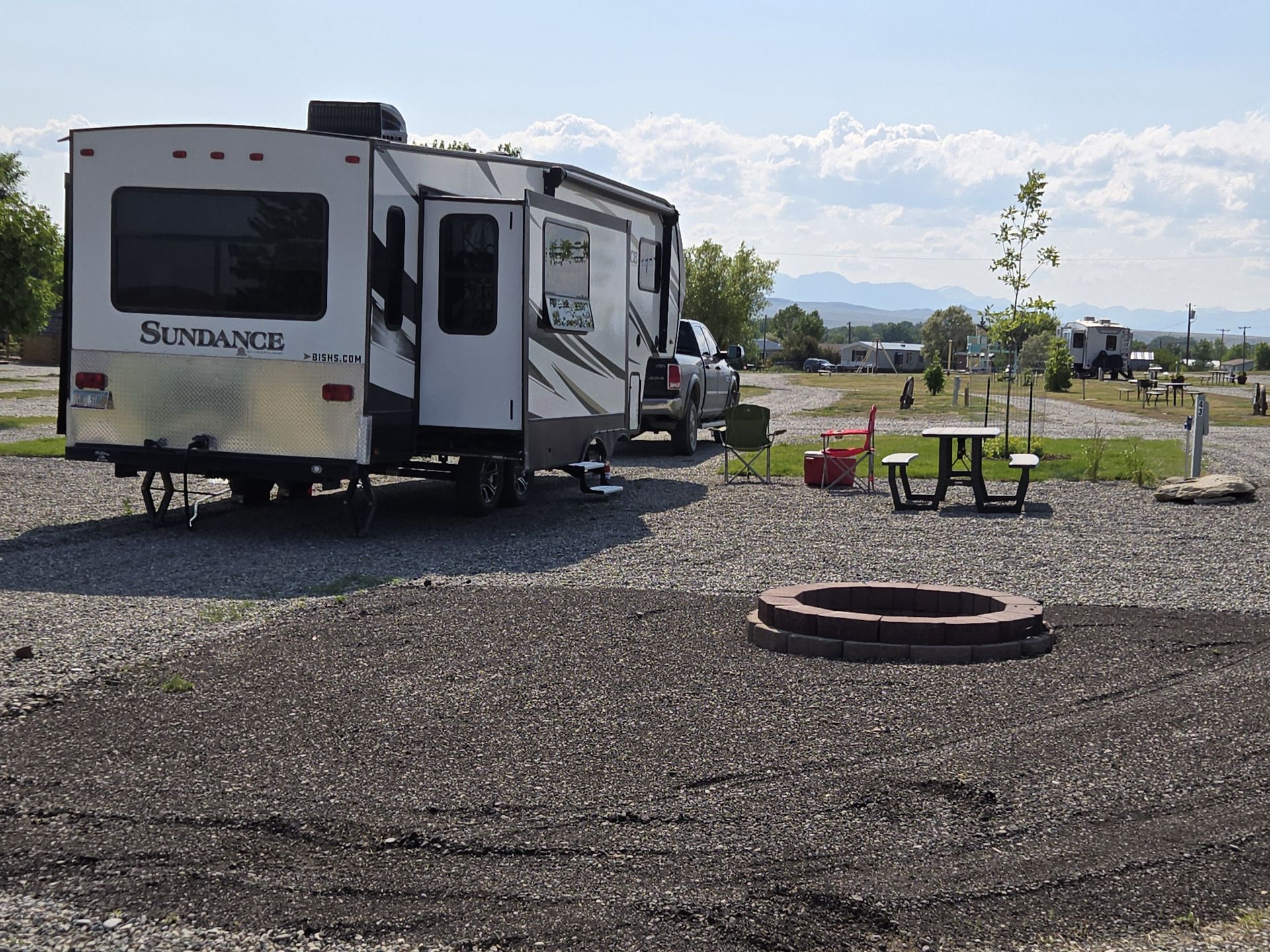 RV parked in gravel campsite with picnic table, fire pit, and distant mountains.
