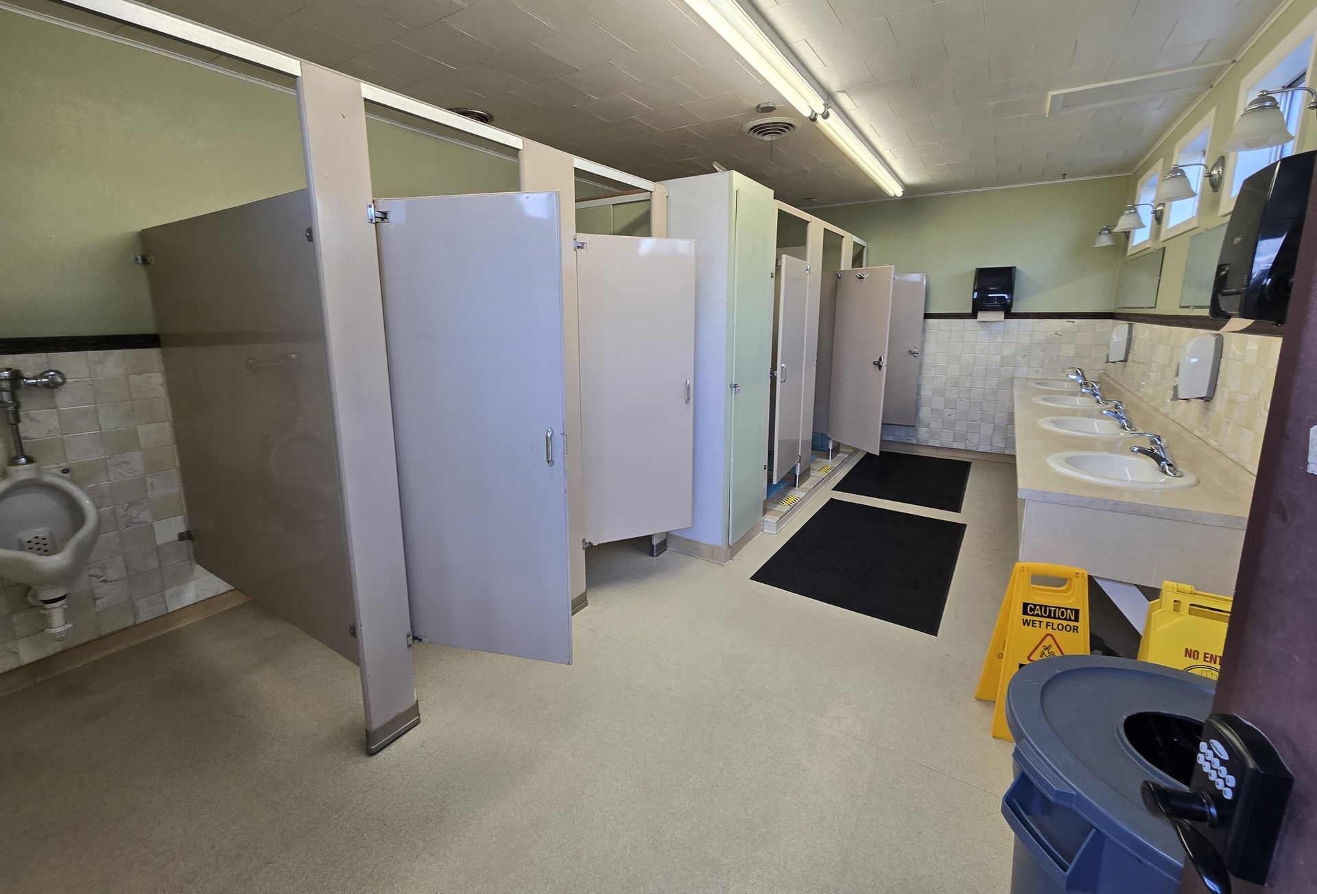 Public restroom with stalls, urinals, sinks, and a trash can. White walls, beige floor, overhead lighting.