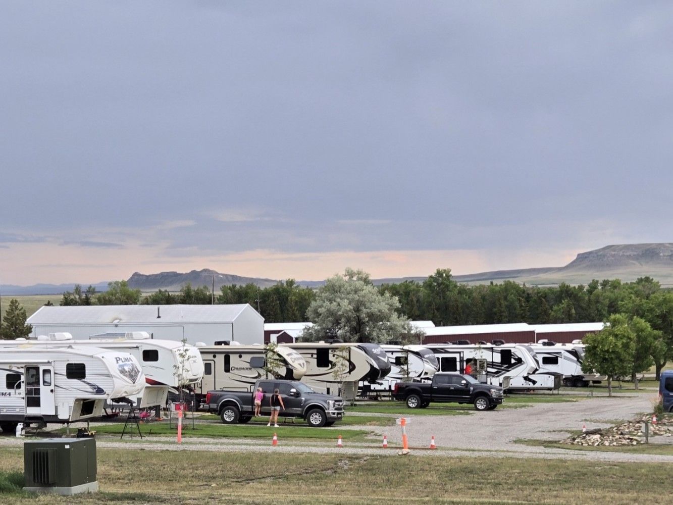 Campground with RVs and trucks, a few people, and mountains in the background under a cloudy sky.
