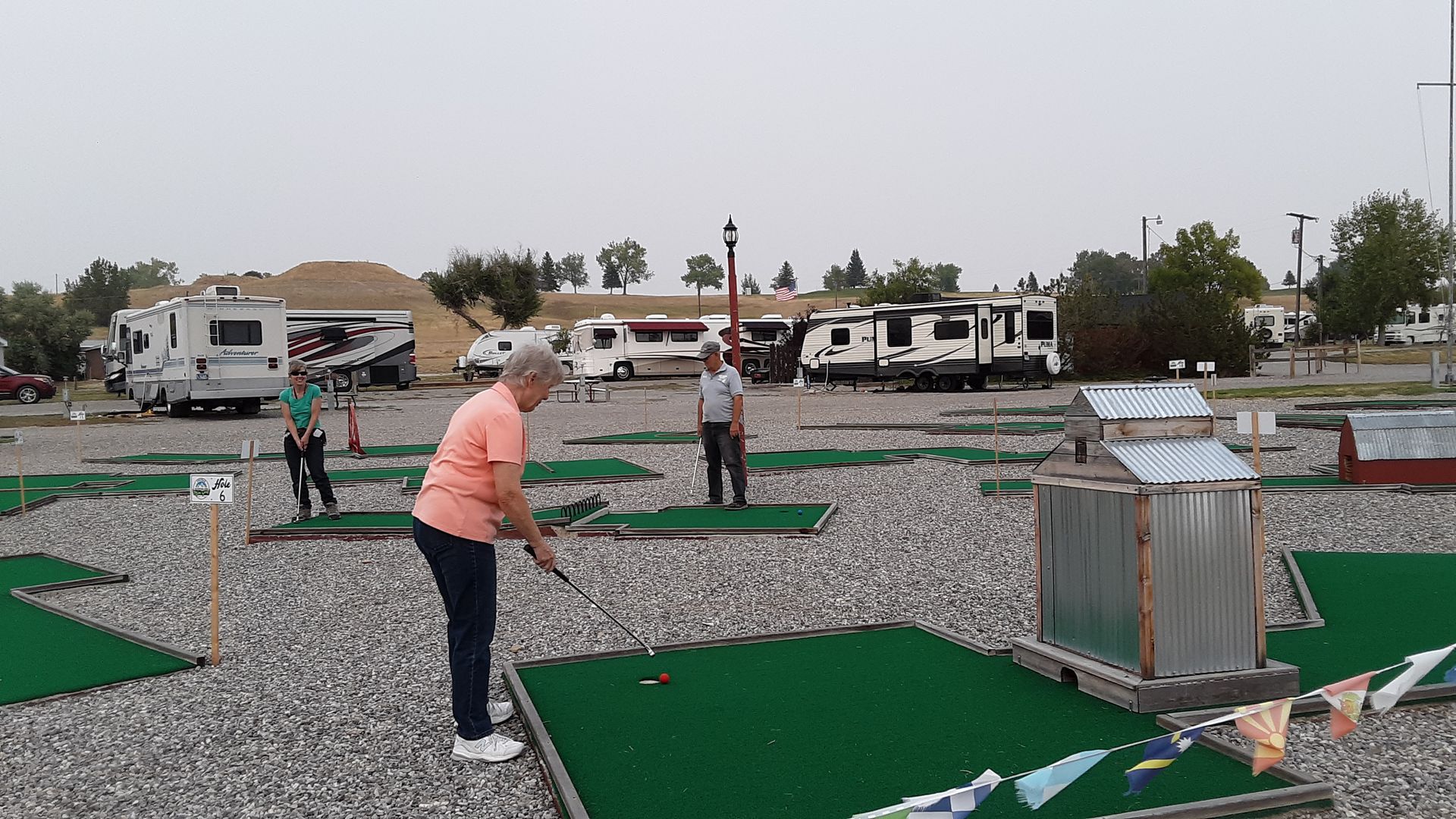 People playing miniature golf outdoors near RVs. A woman in pink shirt swings a club.