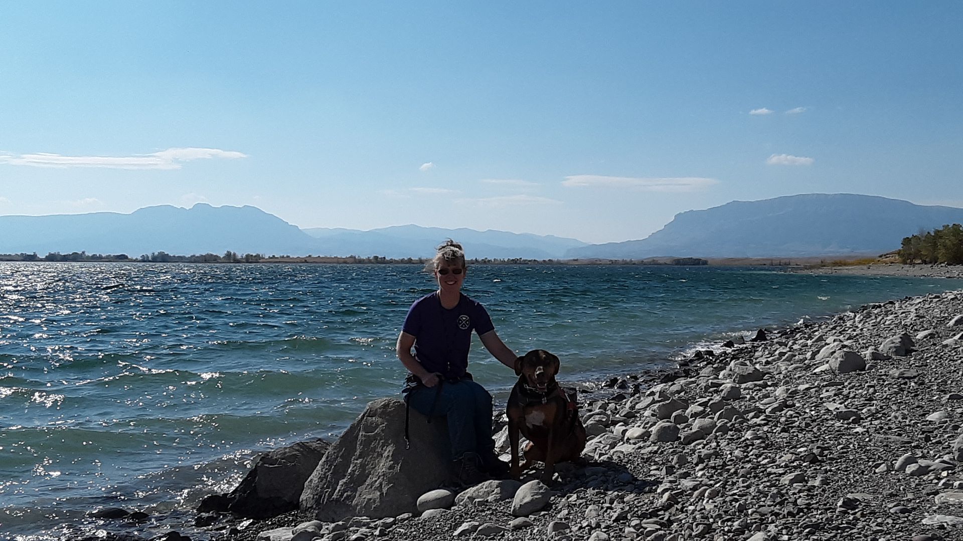 Woman with dog sitting on a rock by a lake with mountains in the background. Sunny day.