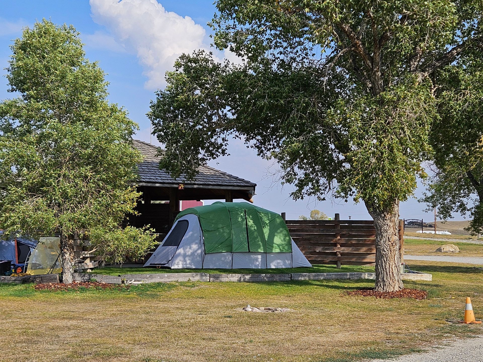 Green and gray tent pitched near trees and a wooden building on a grassy campsite under a blue sky.