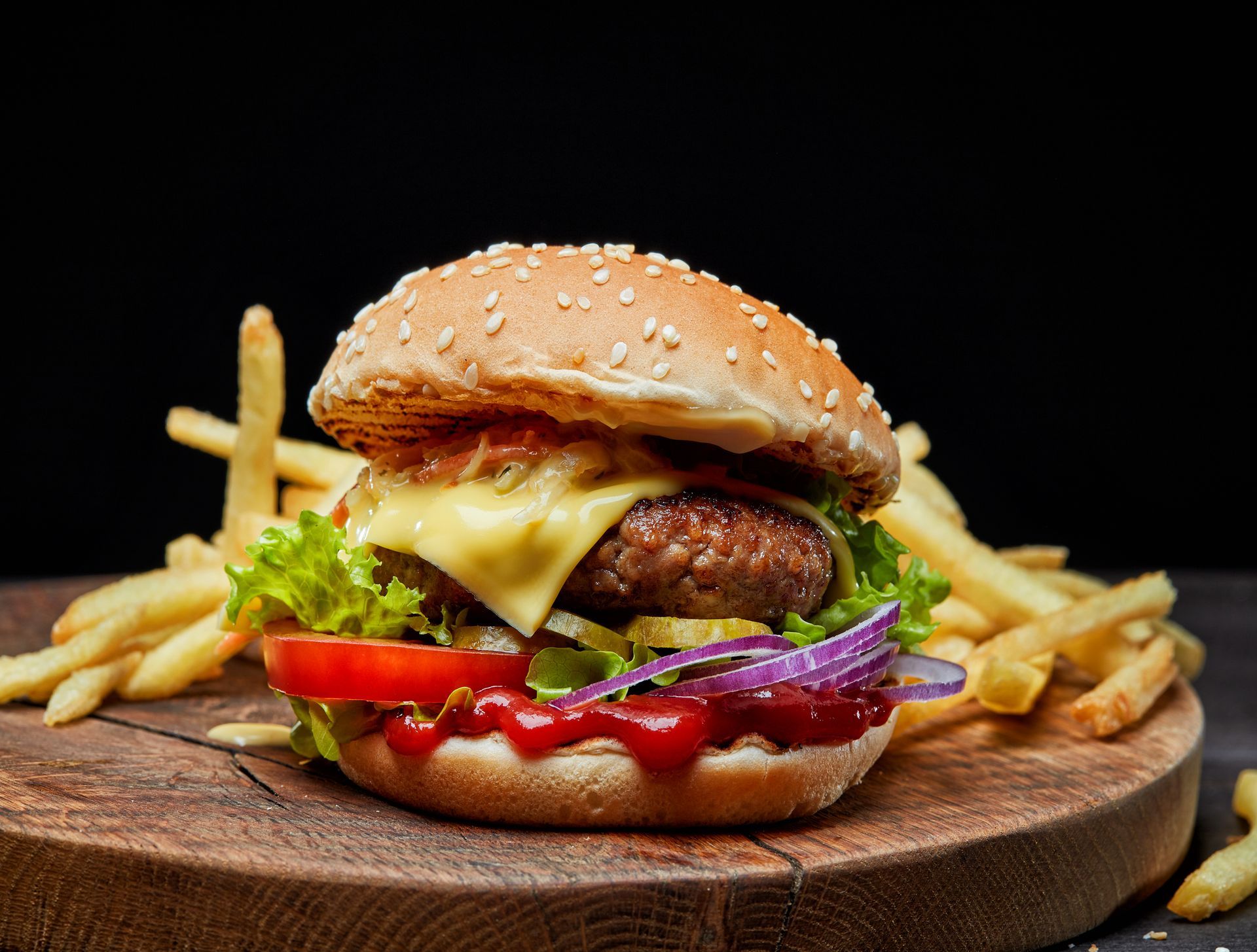 Burger with cheese, lettuce, tomato, and onion on a sesame bun, served with french fries on a wooden board.