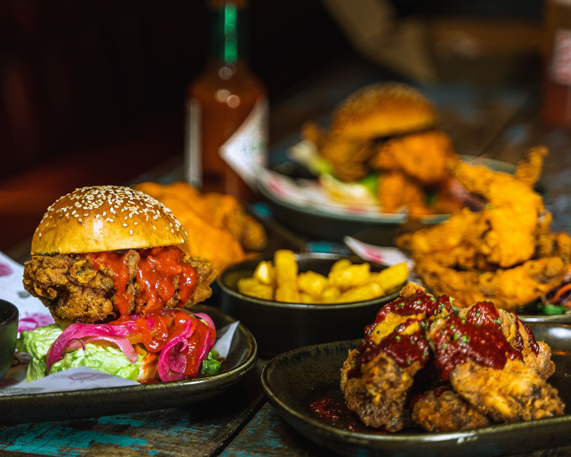 Fried chicken dishes on a table, including a burger, wings, and fries, with a hot sauce bottle in the background.