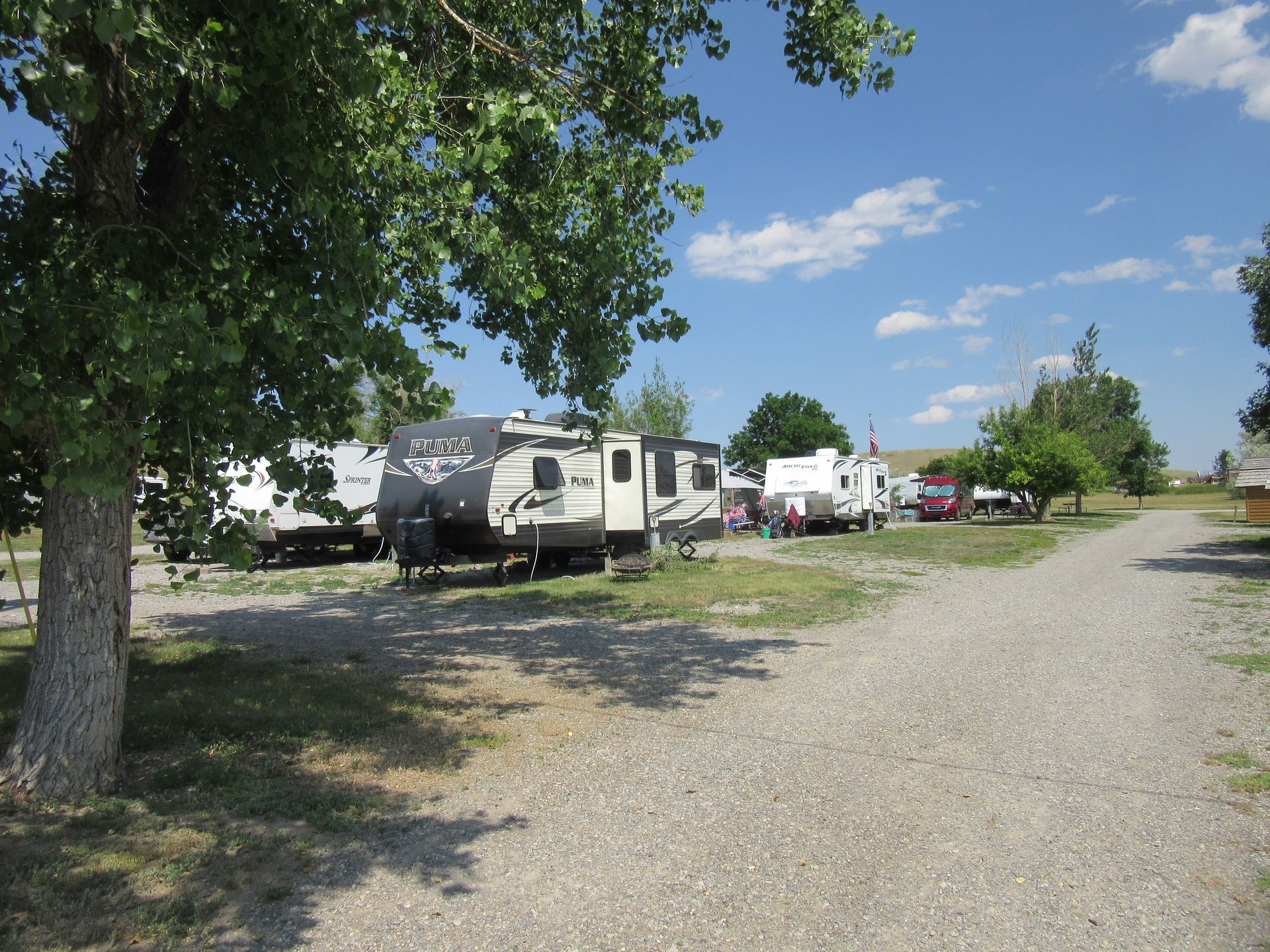 RV campground with gravel paths, RVs parked in spaces, and trees. Blue sky.