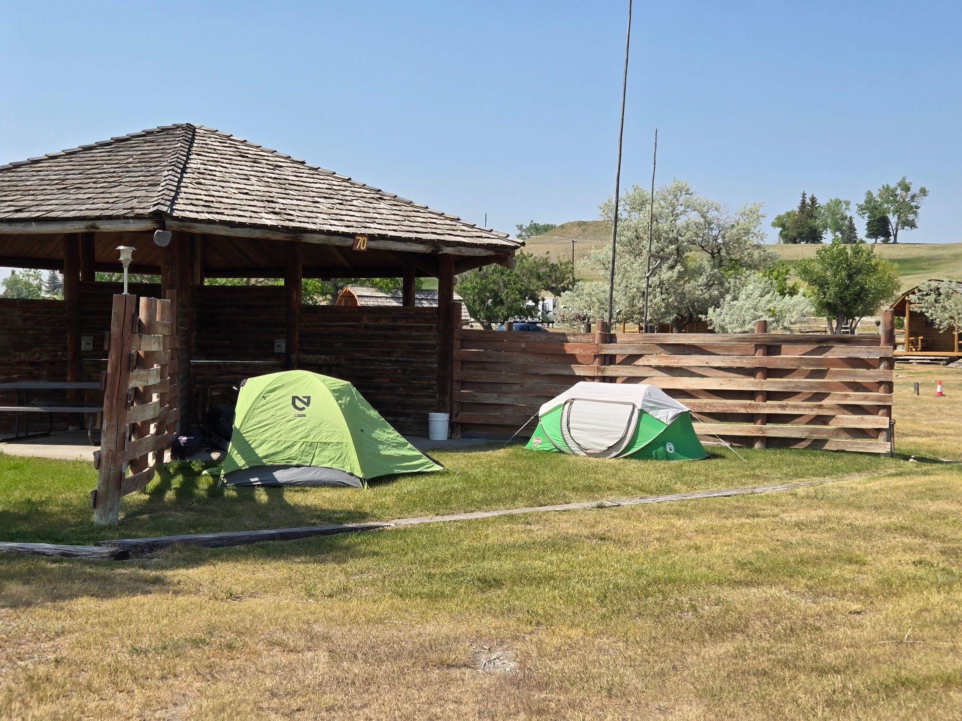 Two tents set up near a wooden gazebo and fence in a grassy area under a blue sky.