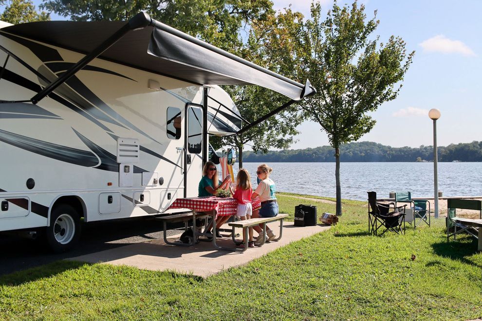 People relaxing beside a white RV under an awning by a lakeside campsite