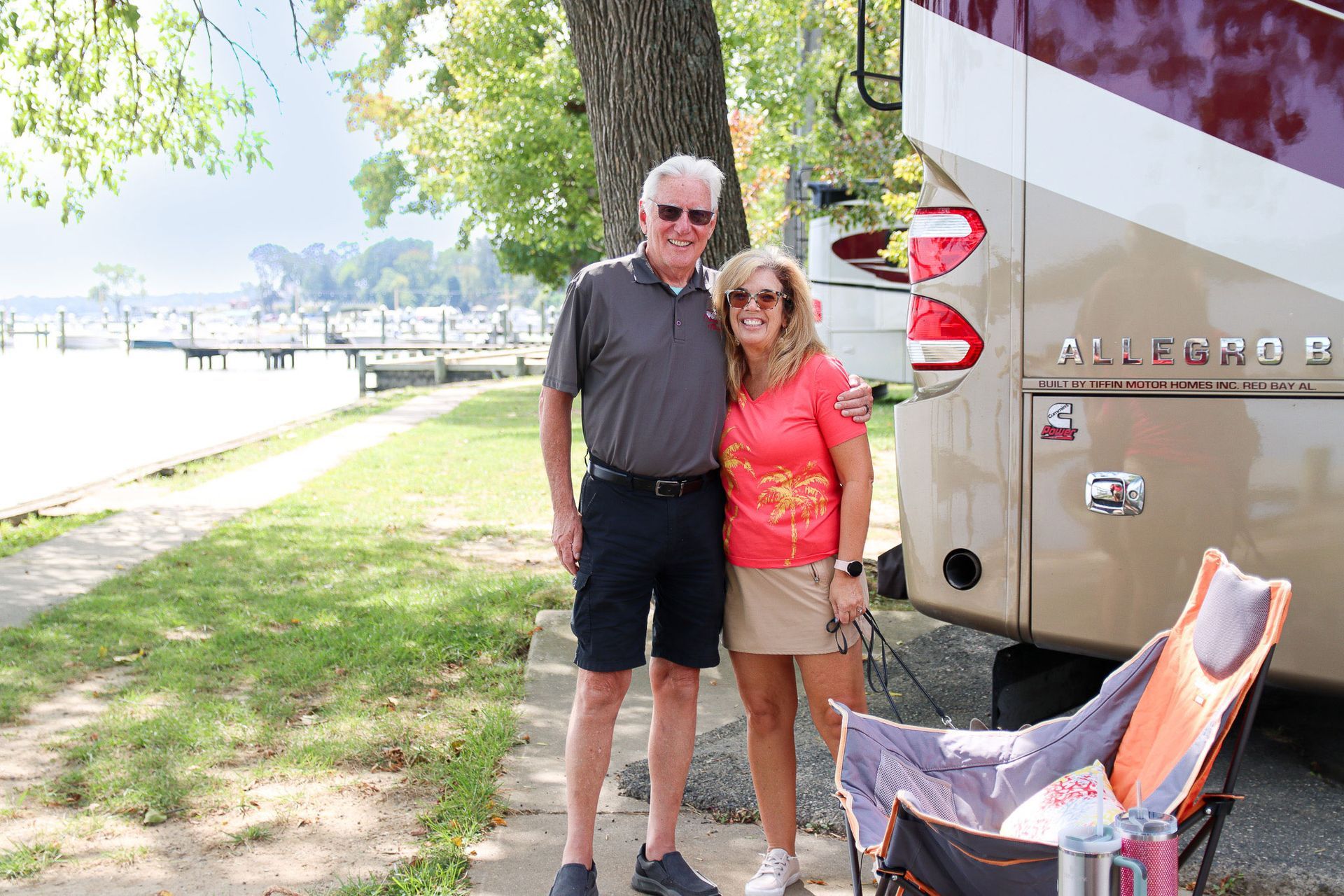 Couple standing beside an RV at a lakeside campsite, with a tree and folding chair nearby.