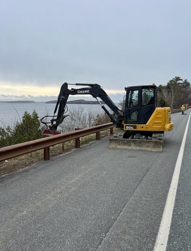 A small excavator is driving down a road next to a body of water.