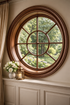 Round wood-framed window above white wainscoting, with flowers and candle on a ledge overlooking green trees