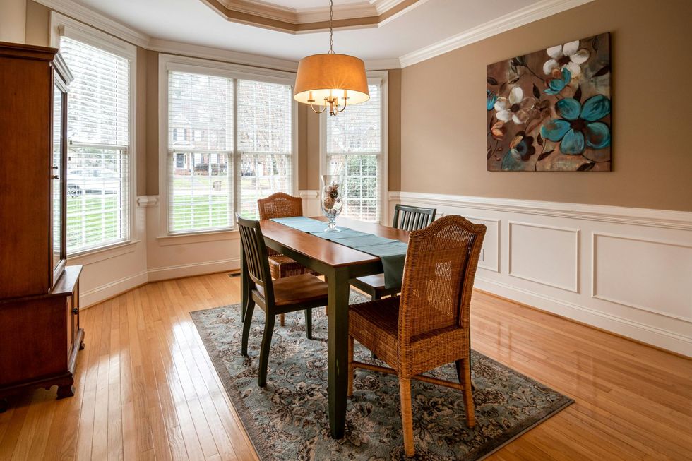 Bright dining room with bay windows, wooden table and chairs, patterned rug, and floral wall art.