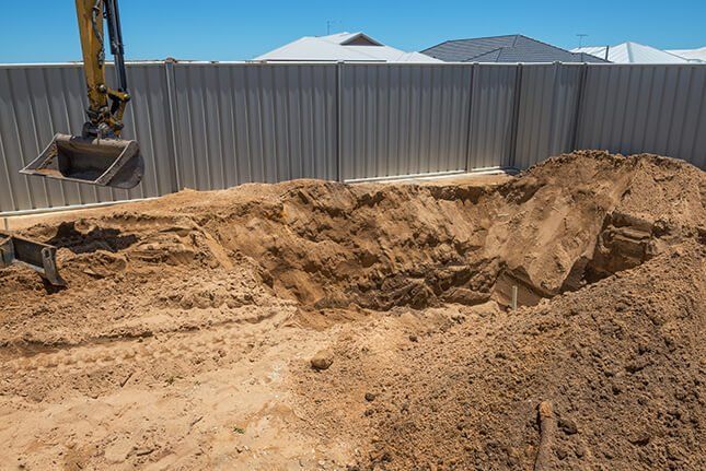 An excavator is digging a hole in the dirt in front of a fence.