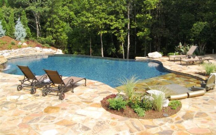 Backyard stone patio with a blue swimming pool, lounge chairs, and trees surrounding it