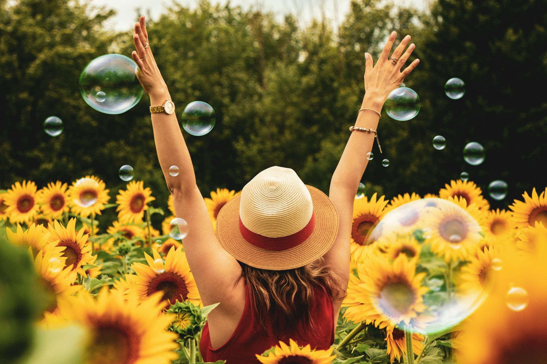 Person in straw hat raising arms in a sunflower field with floating bubbles