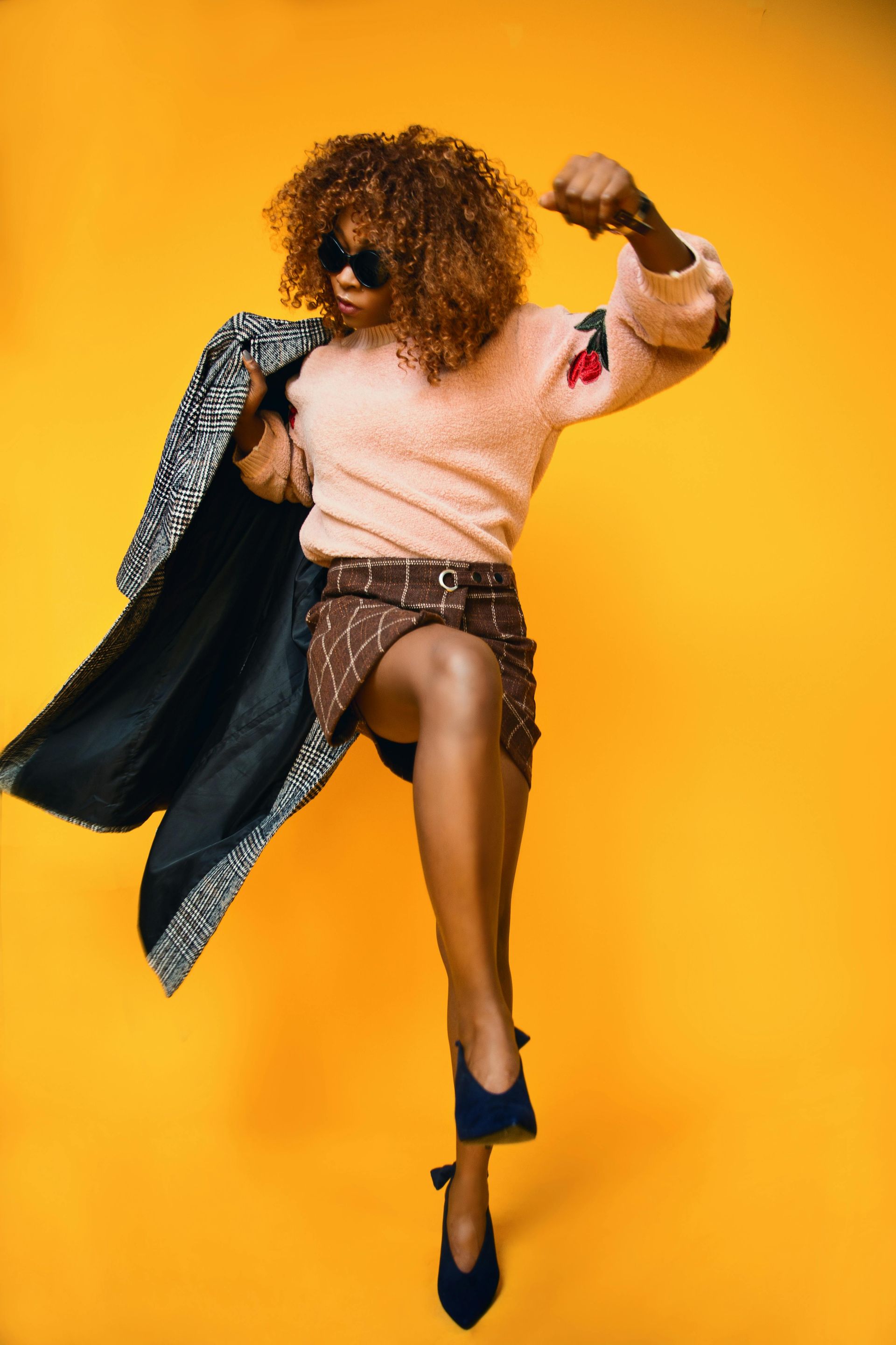 Fashion model jumping in a pink sweater and patterned coat against a bright yellow background